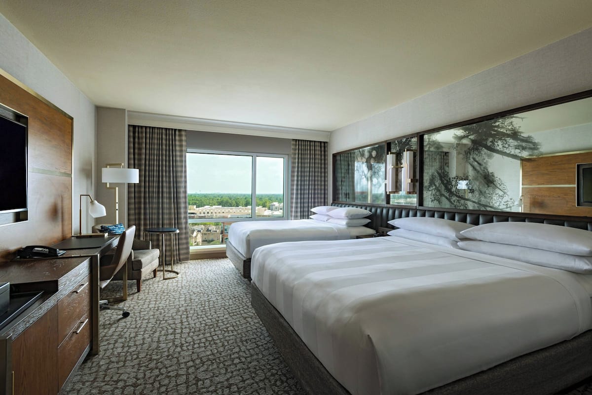 Ein Hotelzimmer mit zwei Betten, einem Schreibtisch, einem Sessel, einem Fernseher und einem großen Fenster mit Blick auf die Stadt.