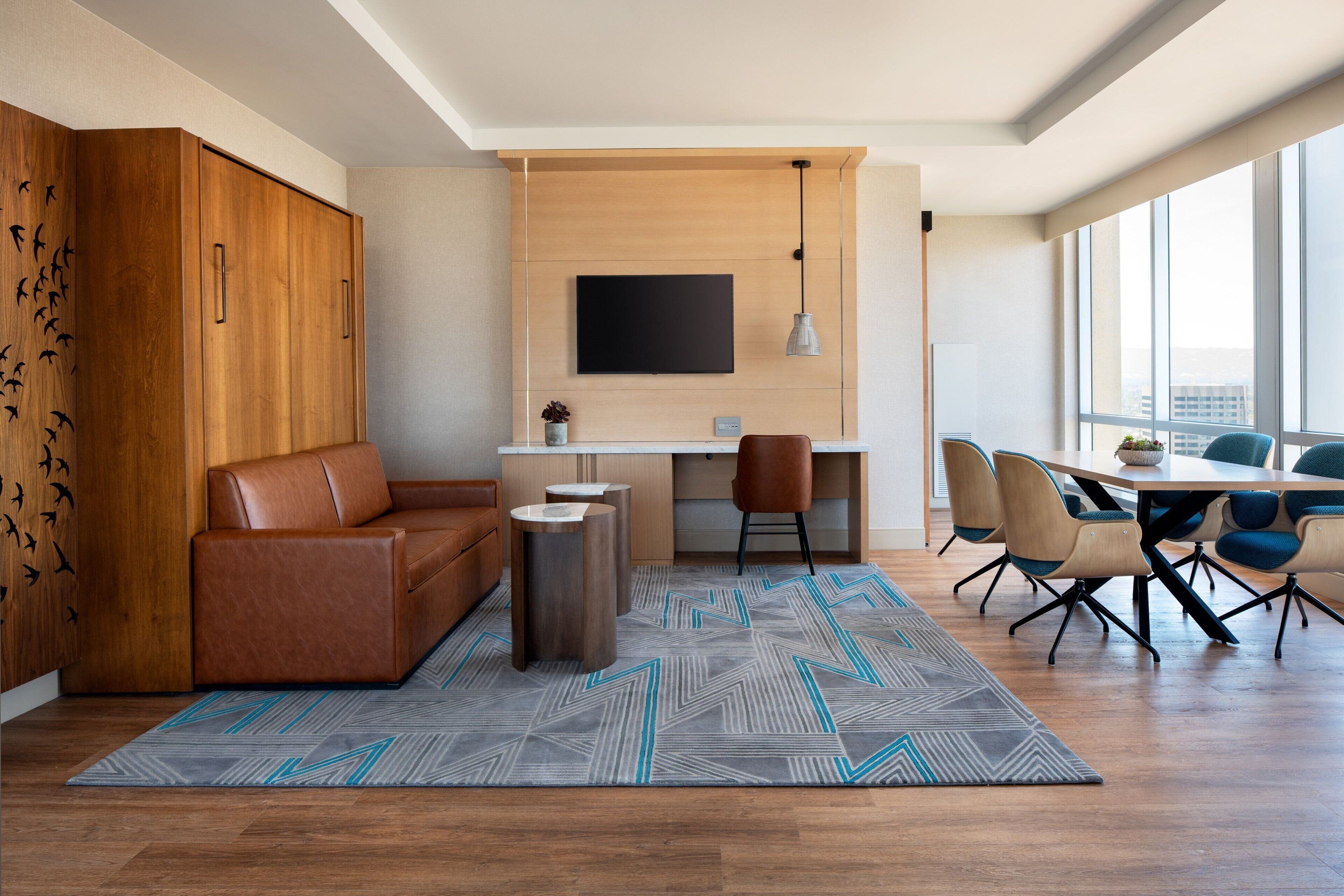 A modern living room with a brown leather sofa, a wooden cabinet, a TV mounted on the wall, a dining area with a table and chairs, and a patterned rug.