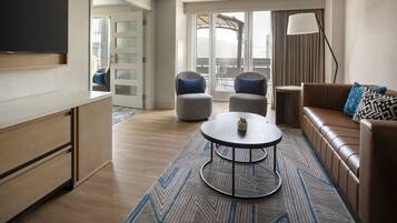 Quarto de hotel moderno com piso de madeira, um sofá, duas poltronas, uma mesa pequena, uma TV e uma varanda com vista.