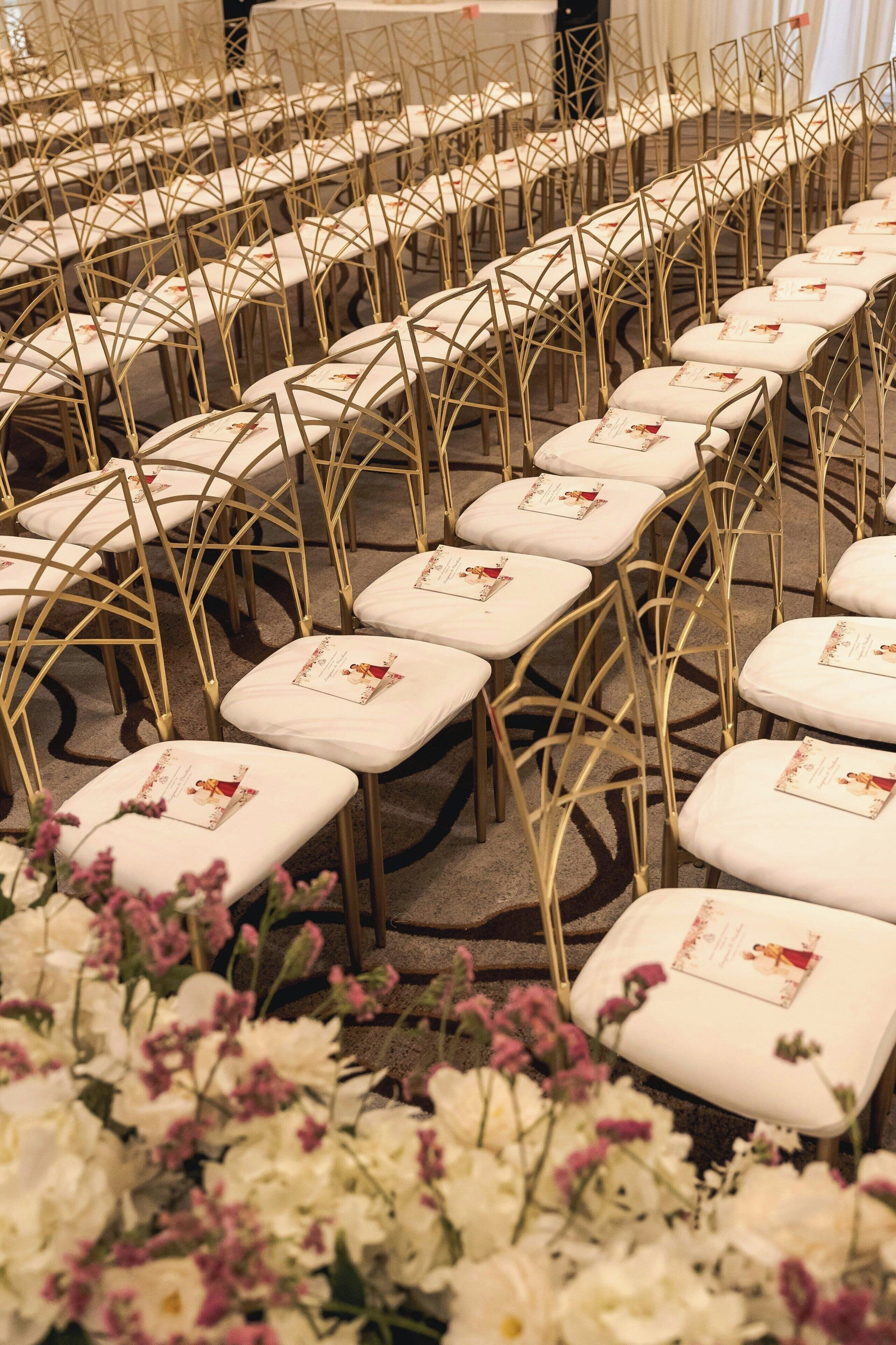 Rows of white cushioned chairs with gold metal frames, arranged in a banquet hall.