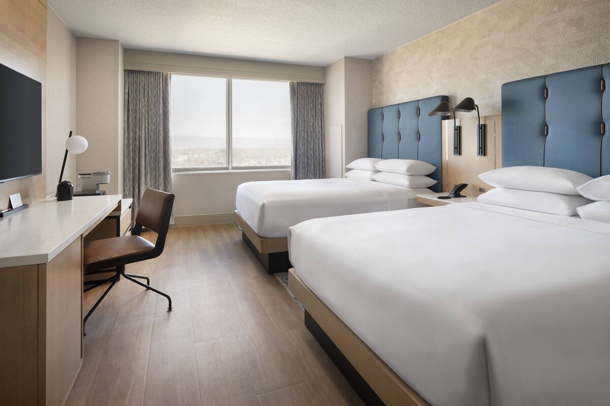 A hotel room with two beds, a desk, a chair, and a large window with curtains.