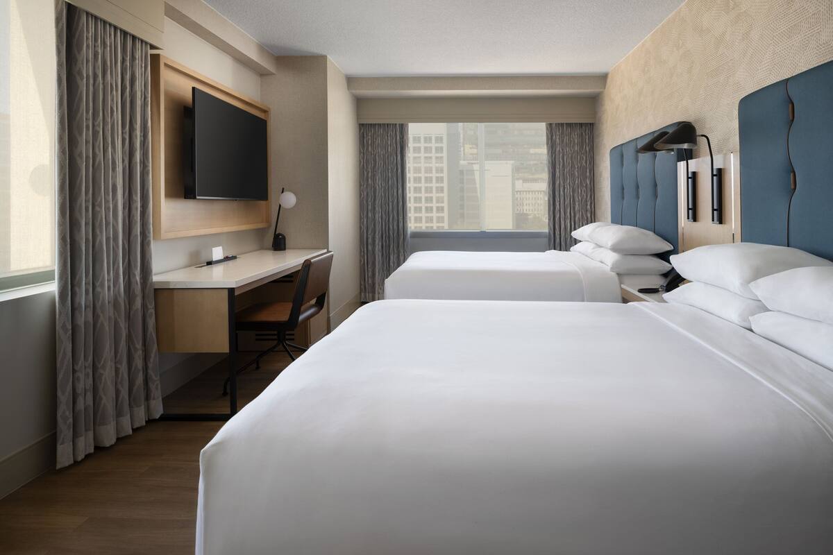A hotel room with a large bed, a desk with a chair, a TV mounted on the wall, and a window with curtains.