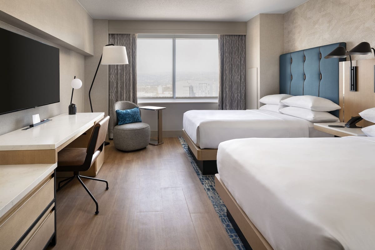A hotel room with a large flat-screen TV, a desk with a chair, two beds, a small round table, and a view of the city through the window.