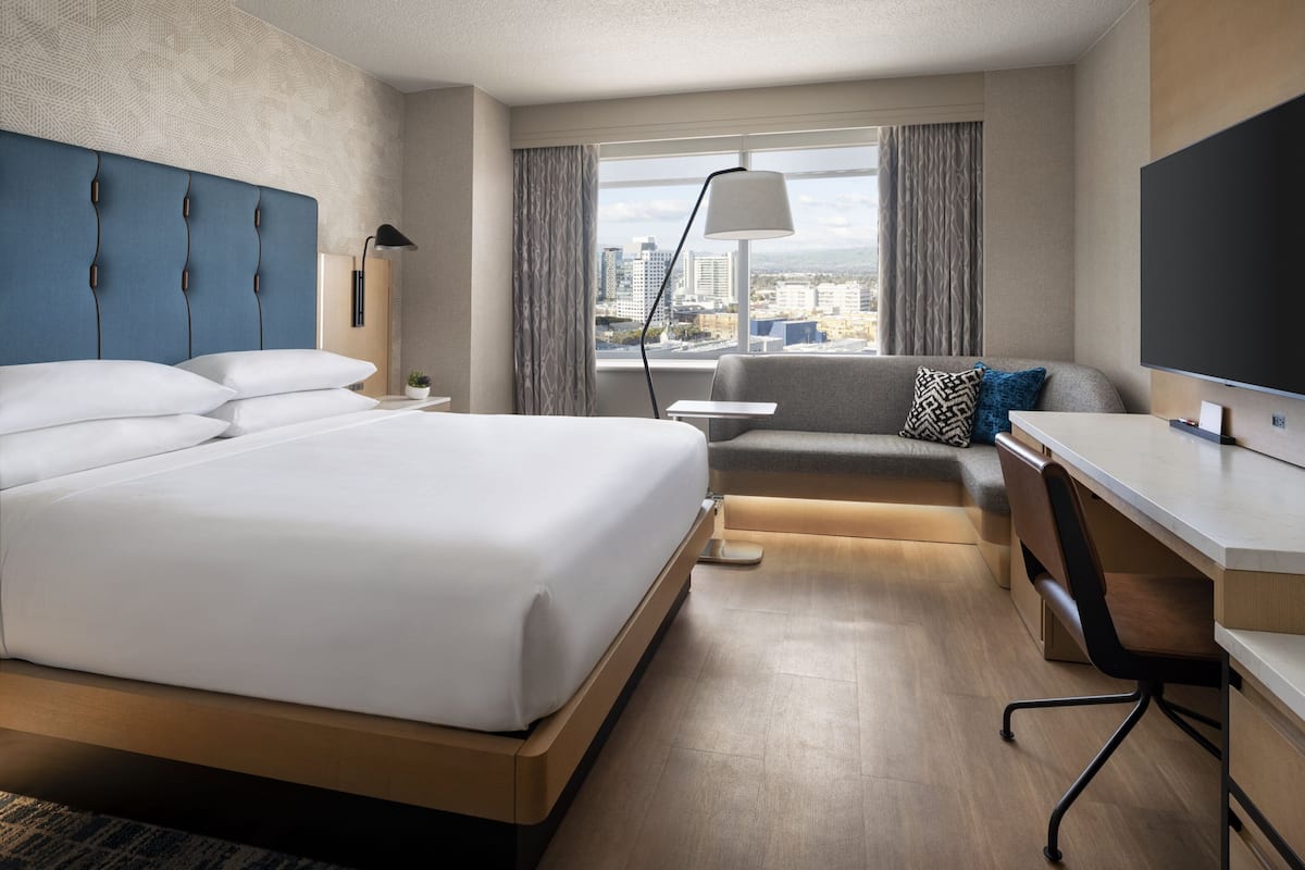A modern hotel room with a large bed, a desk, a sofa, and a city view.