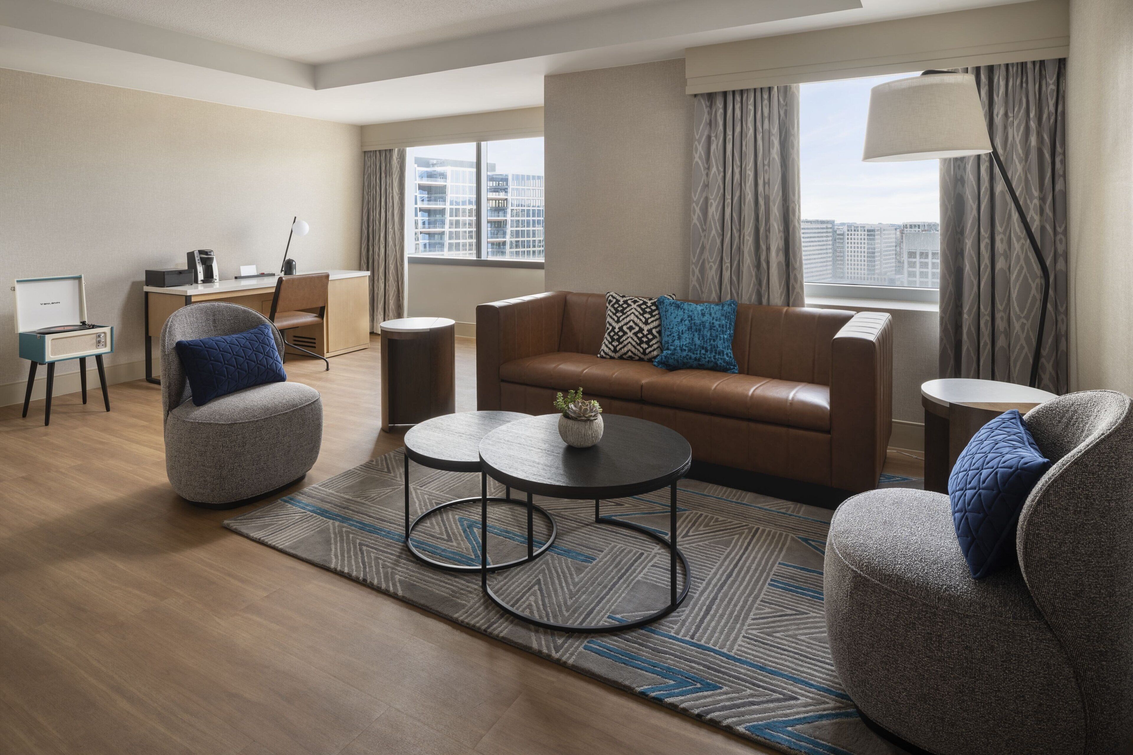 A modern hotel room with a brown leather sofa, two armchairs, a coffee table, a desk, and a view of the city through large windows.