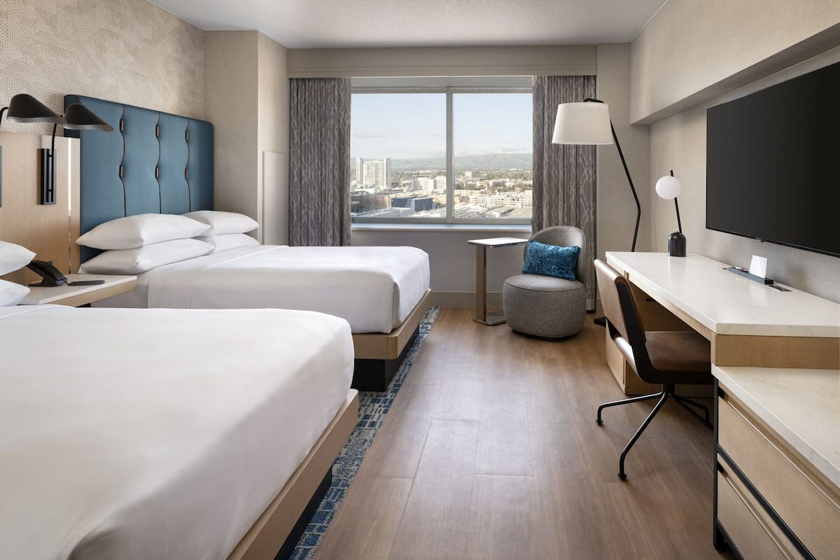 A hotel room with two beds, a desk, a chair, a TV, and a view of the city.