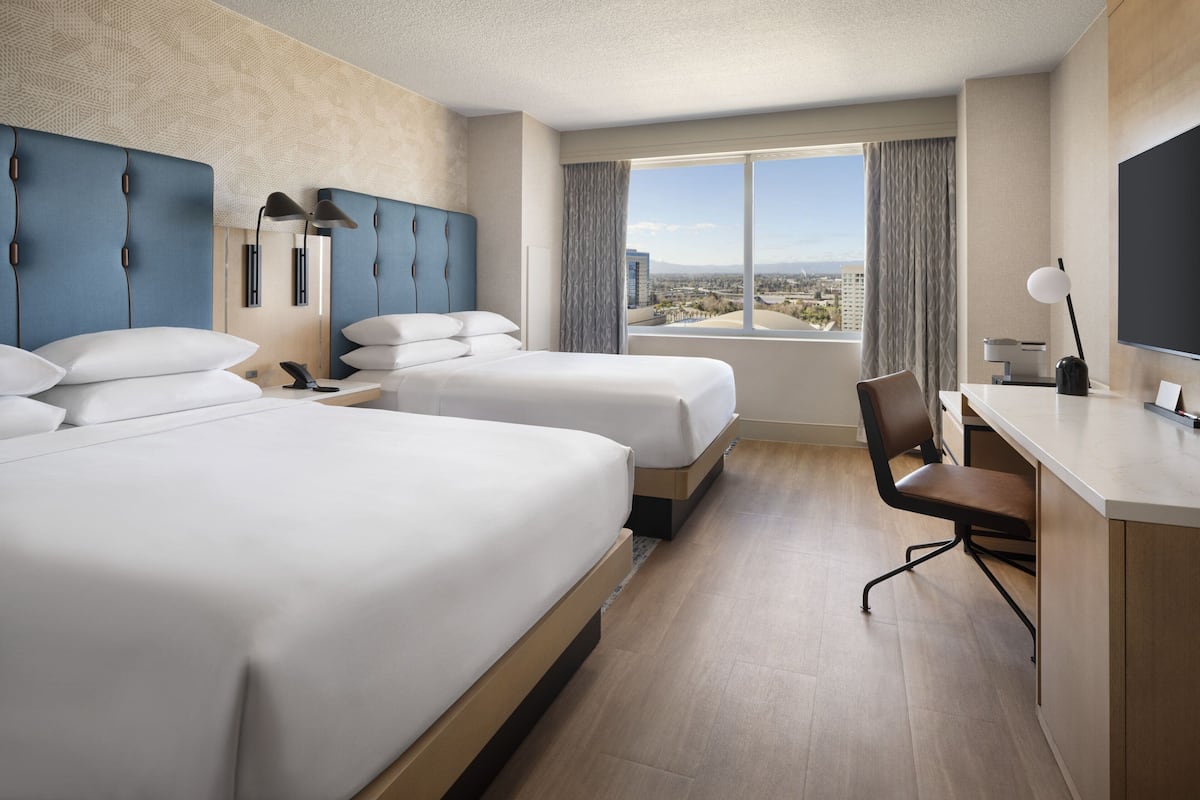 A hotel room with two beds, a desk, a chair, a TV, and a large window offering a city view.