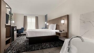 A modern hotel room with a bed, a desk, a chair, a desk lamp, a nightstand, a wall-mounted TV, and a bathroom with a bathtub and a mirror.