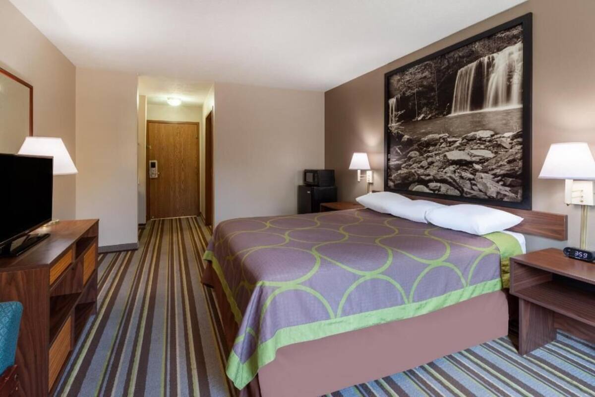 A hotel room with a bed, a television, a desk, a chair, a lamp, a clock, and a painting of a waterfall.