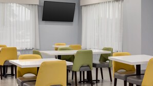 Un salón de conferencias con mesas y sillas, un televisor de pantalla plana en la pared y grandes ventanales con cortinas translúcidas.