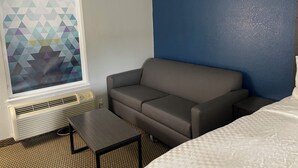 A hotel room with a gray sofa, a small table, a bed with a white quilt, a window with a geometric pattern, and a blue accent wall. - Clairmont Inn & Suites Warren (Warren)