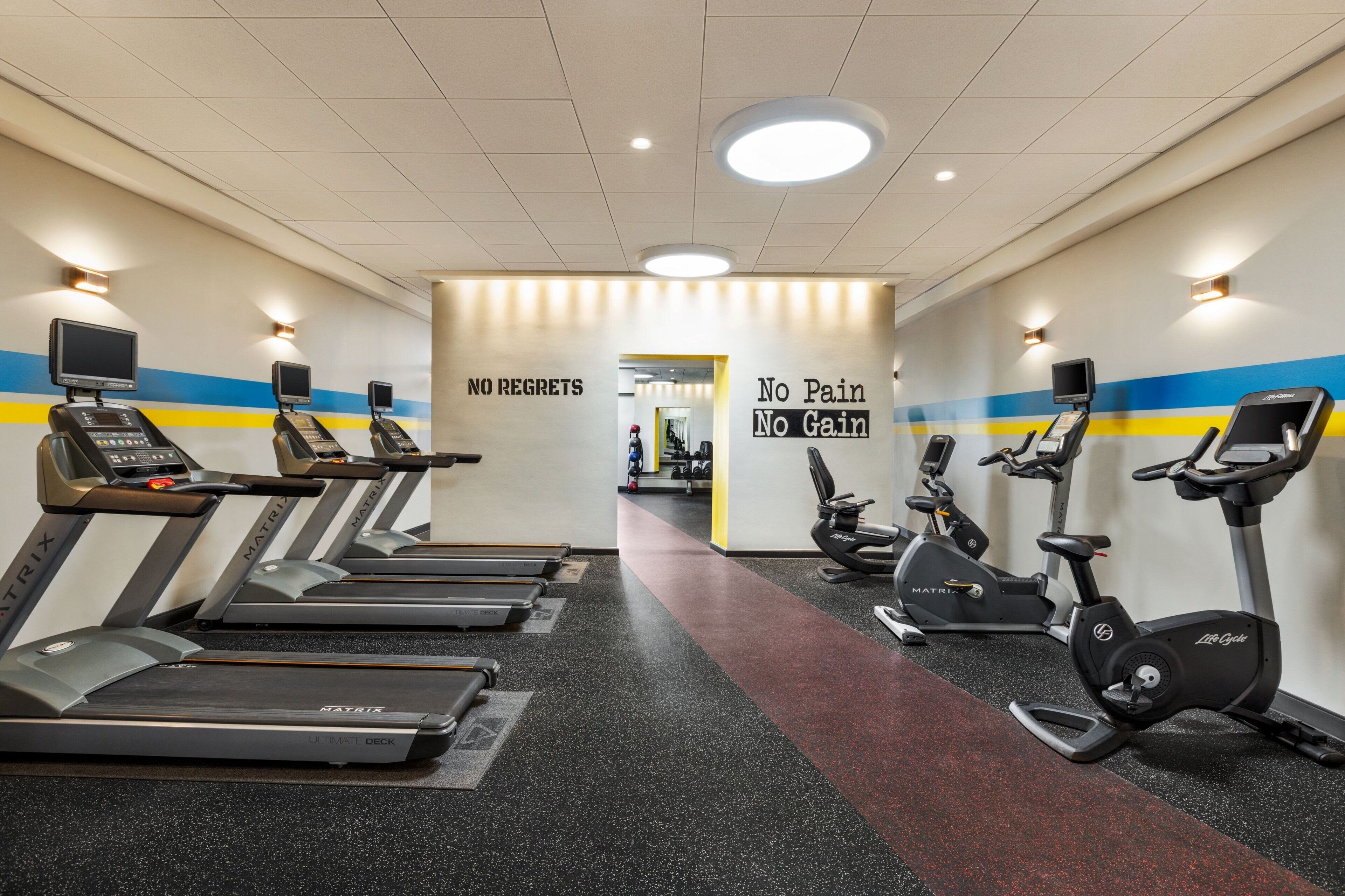 A gym with treadmills, stationary bikes, and a motivational wall with phrases like "NO REGRETS" and "No Pain No Gain".
