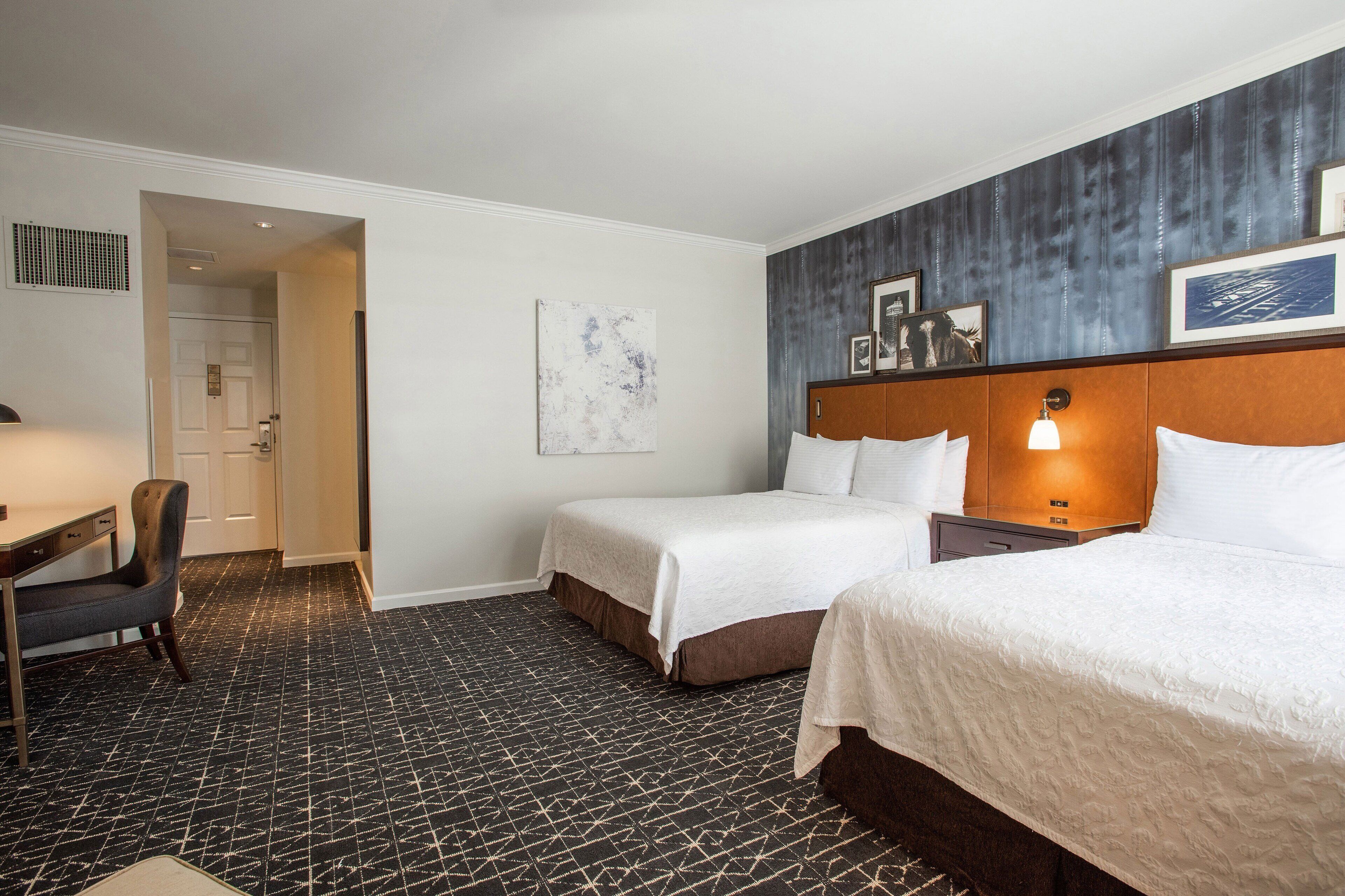 A hotel room with two beds, a desk, and a chair. There is a painting on the wall and a picture frame above the bed.