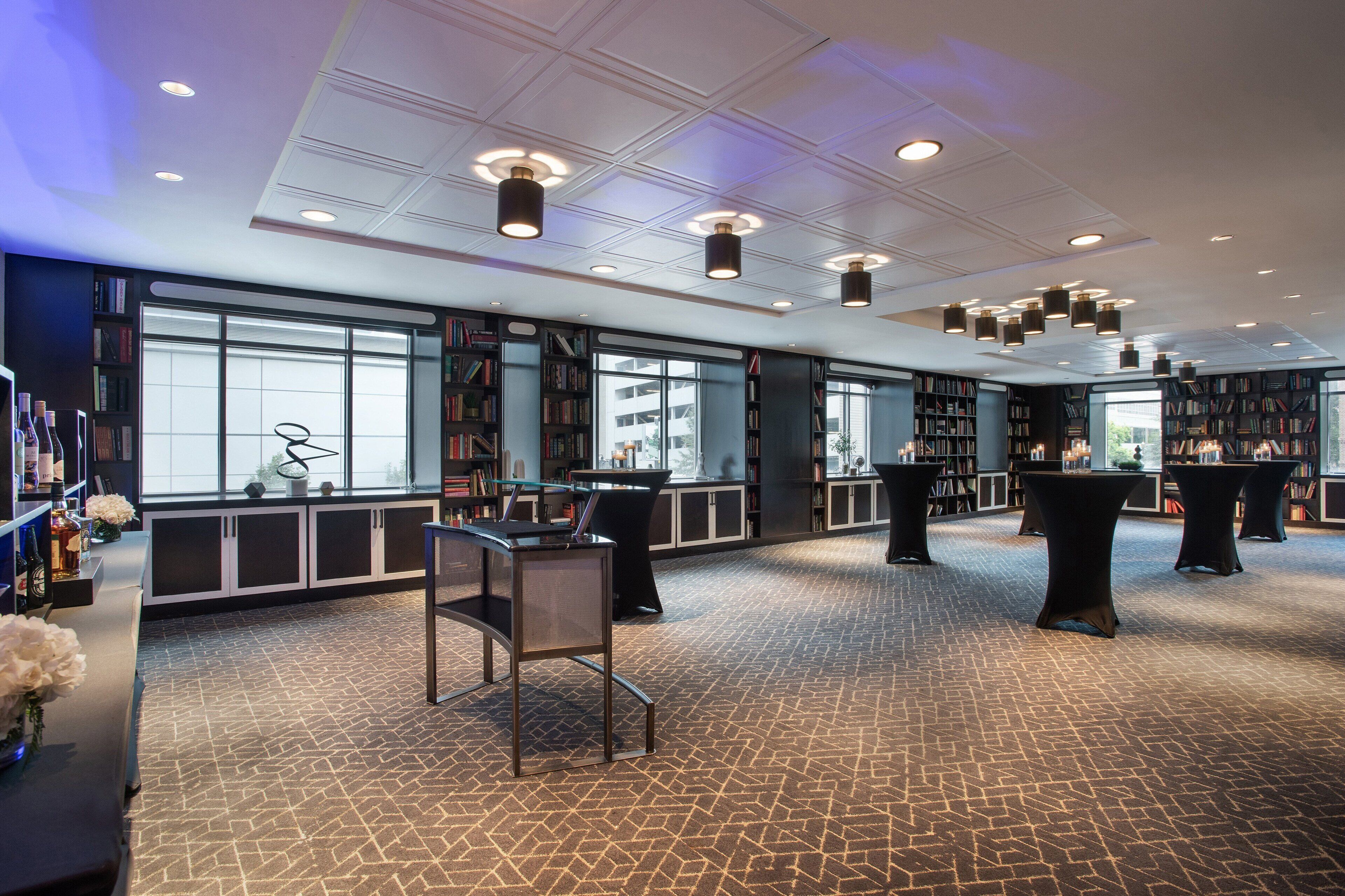 A spacious library with bookshelves, tables, and chairs, featuring large windows and a patterned floor.