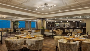 A spacious restaurant with high ceilings, multiple dining tables, wicker chairs, and a bar area with a large selection of bottles.