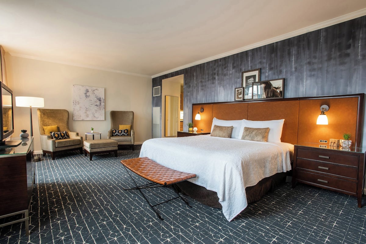 Ein Hotelzimmer mit einem großen Bett, einem kleinen Schreibtisch, einem Stuhl, einer Bank und einem Schminktisch.