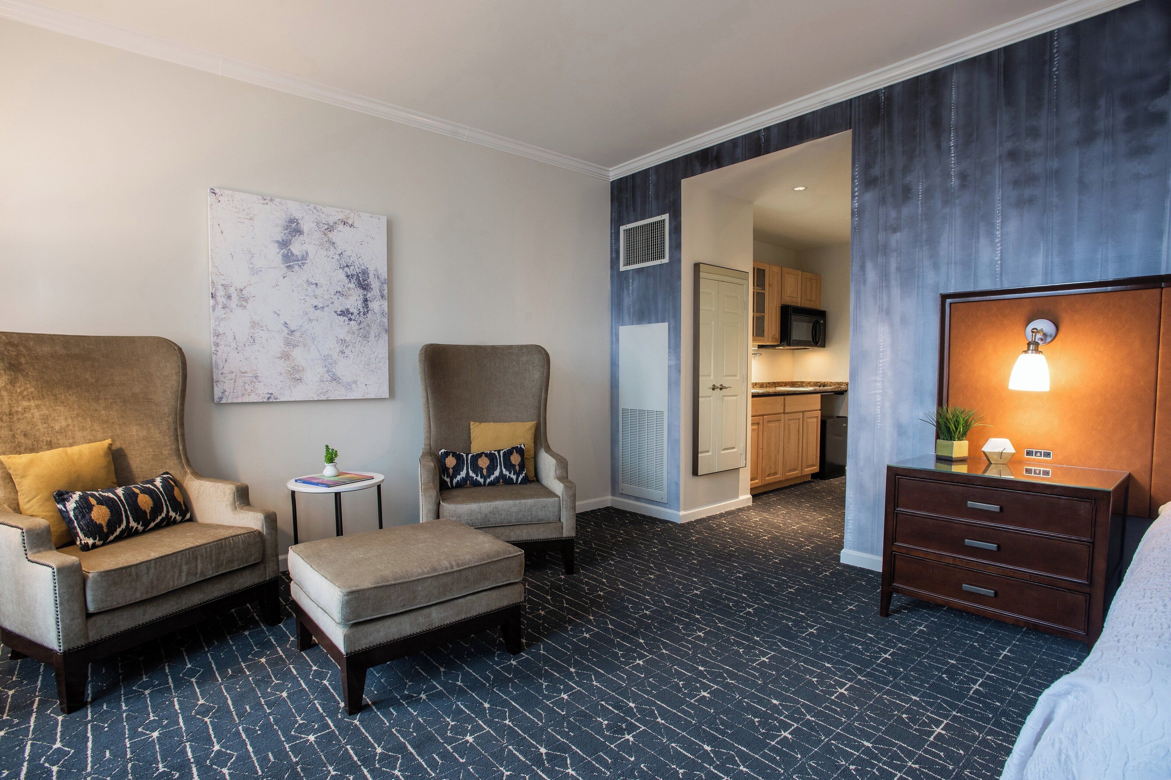A hotel room with a sofa, armchair, ottoman, a small side table with a plant, a cabinet, and a large painting on the wall.