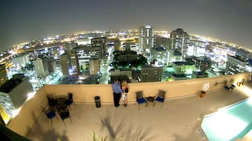 A rooftop terrace with a pool, chairs, and a view of a cityscape at night.