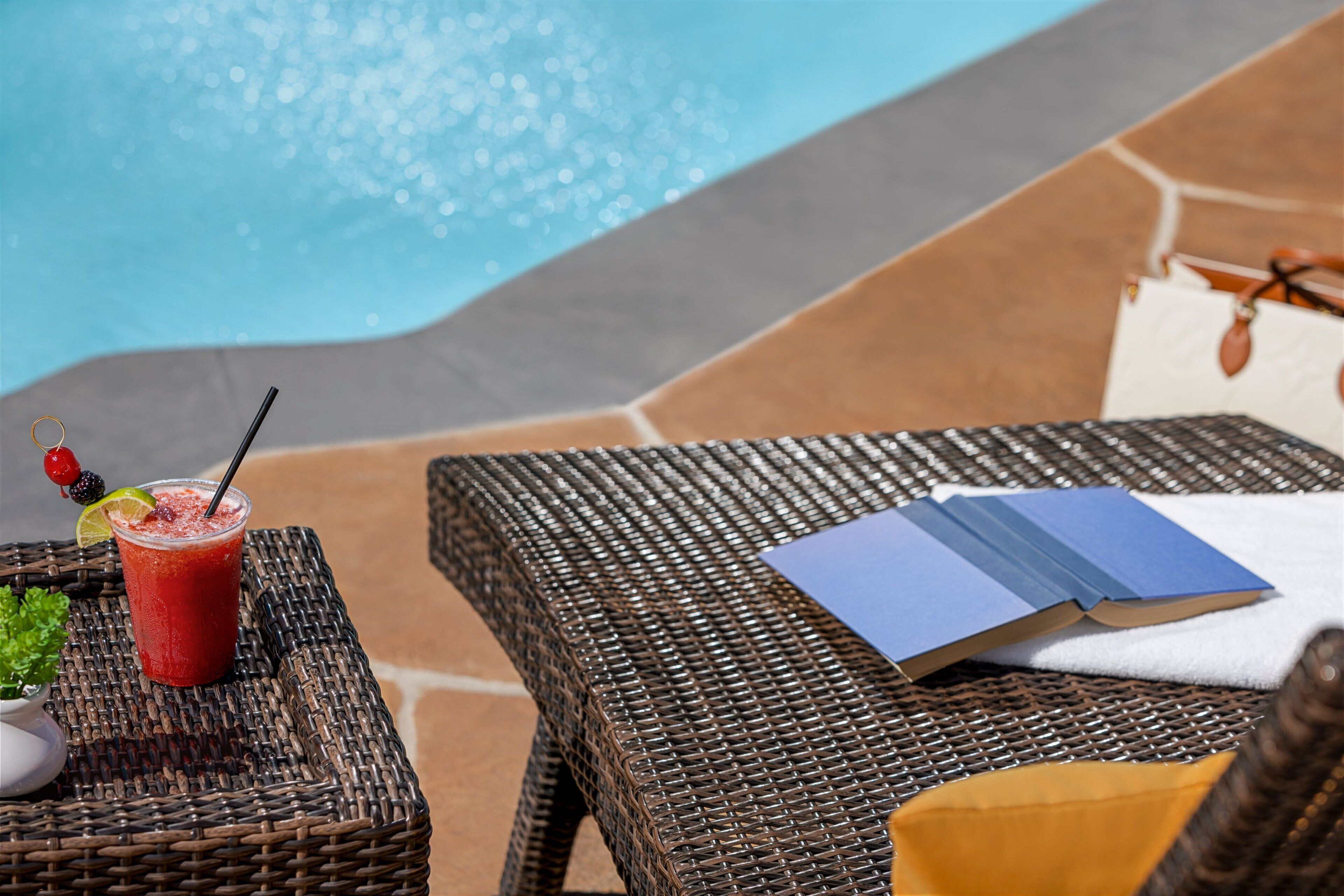 A wicker lounge chair with a drink and a book beside a pool.