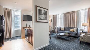 Una habitación de hotel moderna con cocina, sala de estar con sofá y mesa de centro, y un ventanal con cortinas.