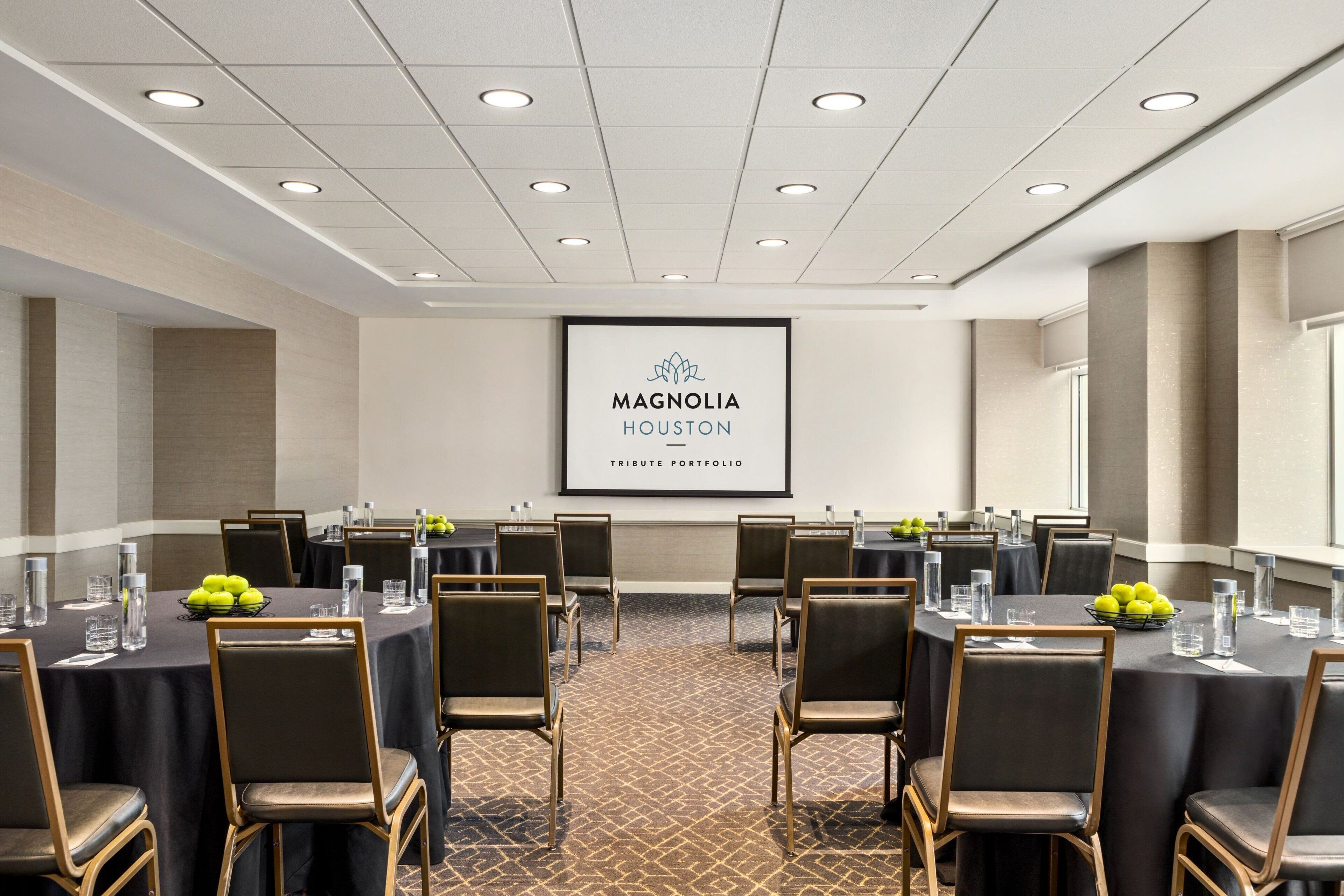 Un salón de conferencias con mesas redondas, sillas y una pantalla proyectora mostrando "Magnolia Houston Tribute Portfolio".