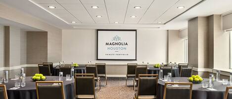 Un salón de conferencias con mesas redondas, sillas y una pantalla proyectora mostrando "Magnolia Houston Tribute Portfolio".