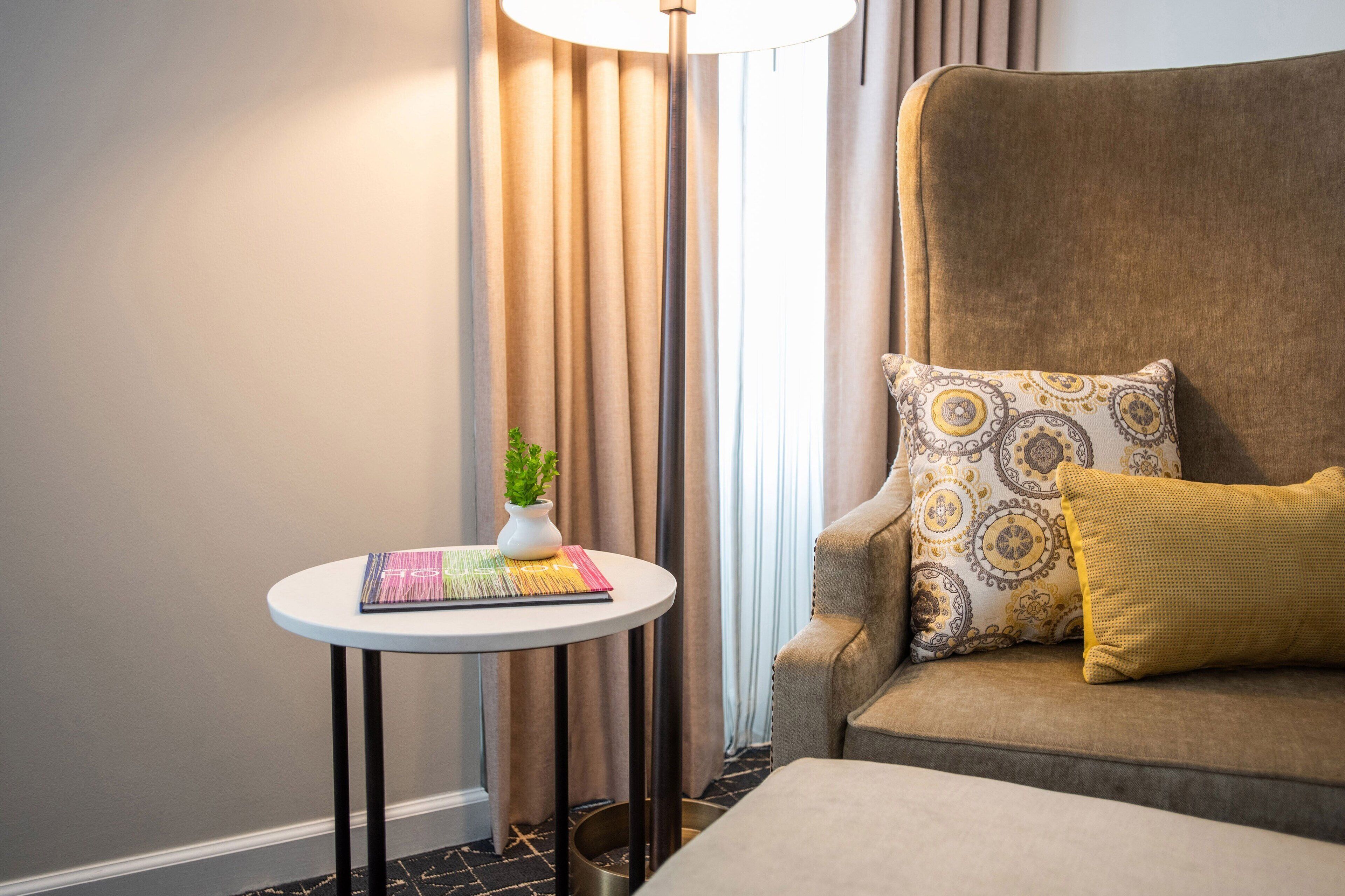 A cozy room with a brown armchair, a small round table with a plant and a colorful mat, and a lamp.