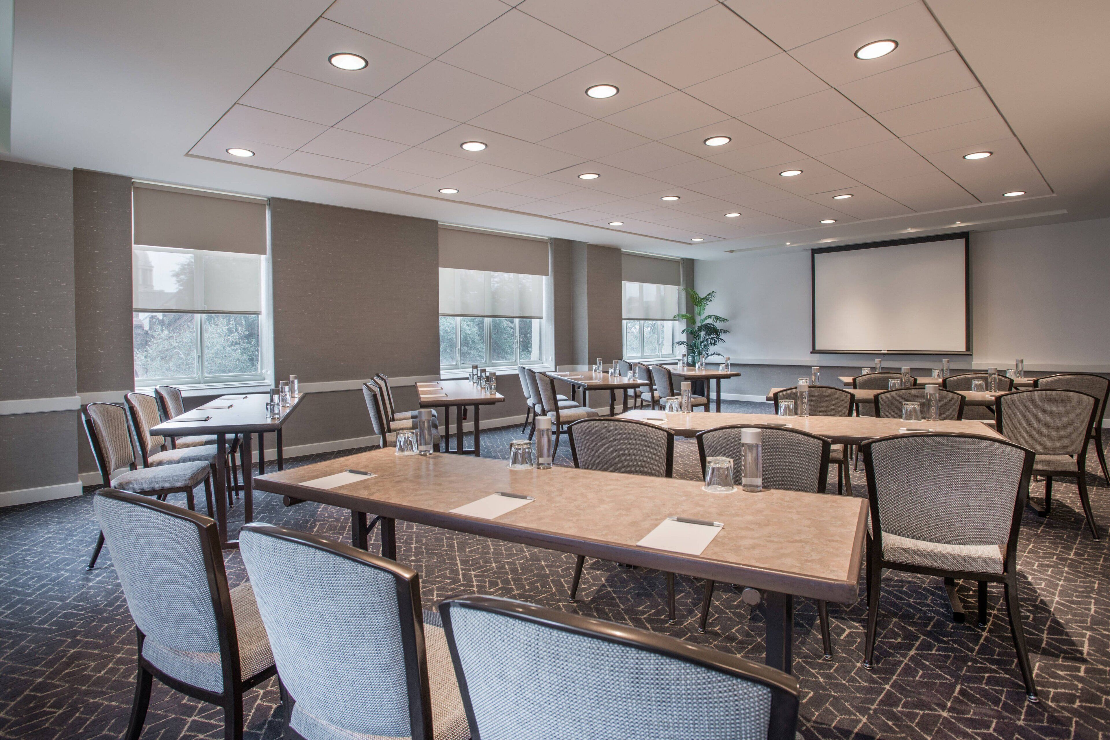 A conference room with multiple tables and chairs, large windows, and a projector screen.