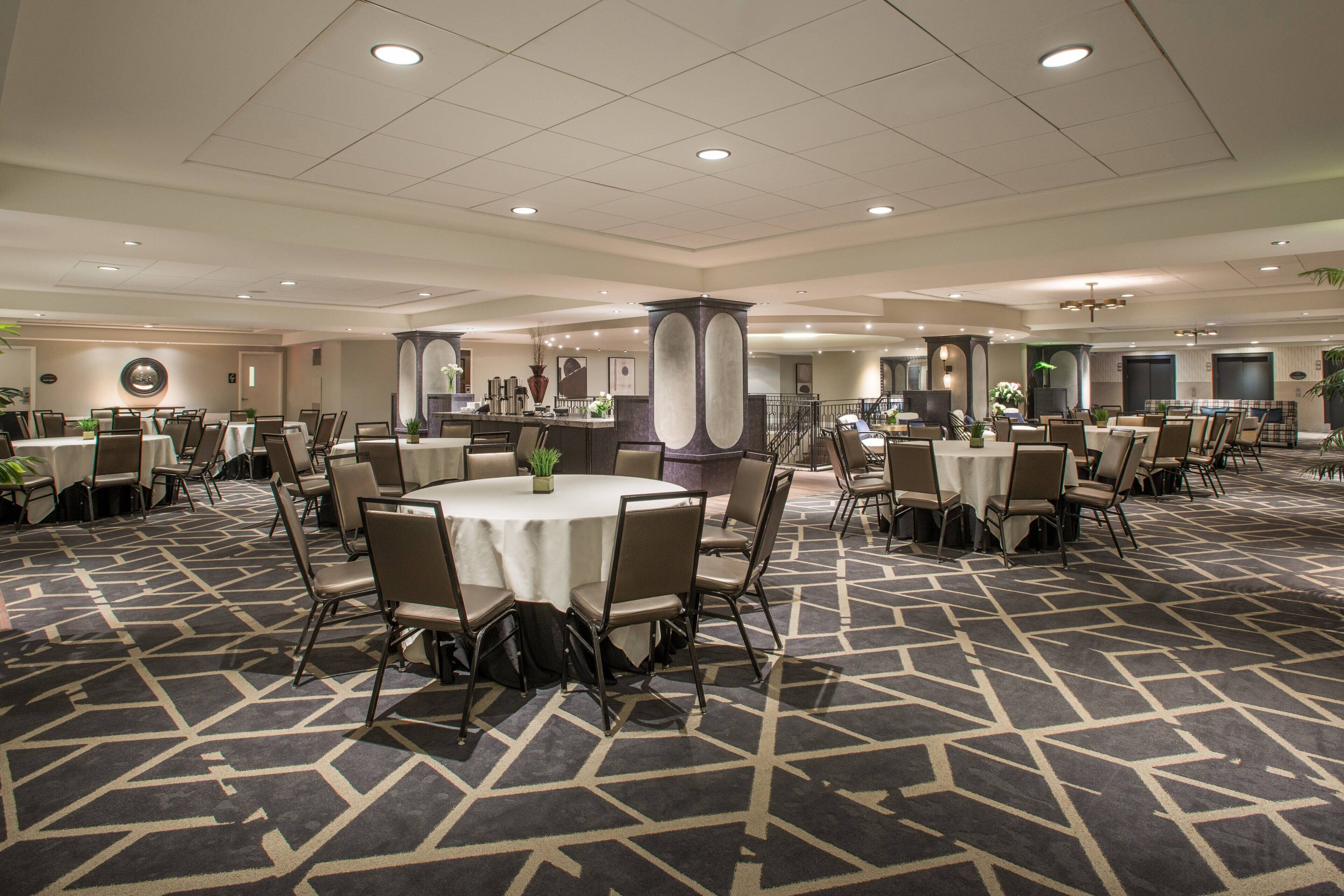 A spacious hotel lobby with multiple round tables and chairs, a patterned carpet, and a modern ceiling design.