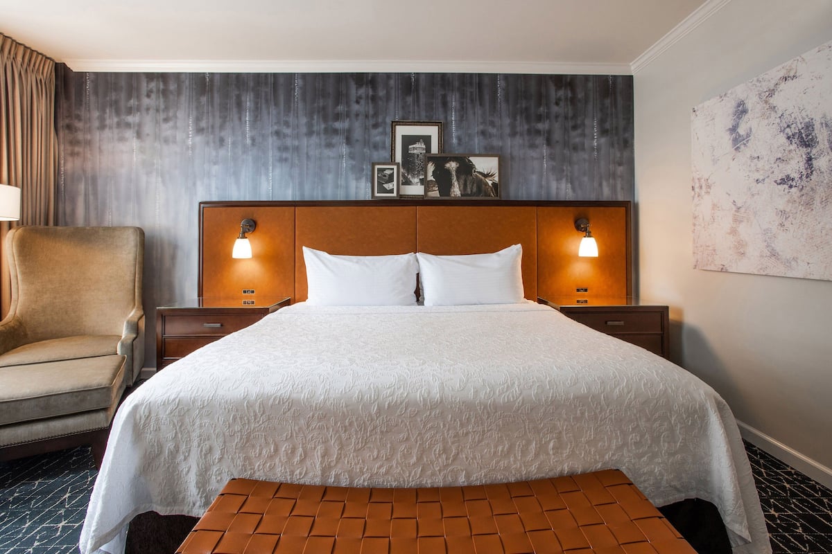 Ein Hotelzimmer mit einem großen Bett, zwei Nachttischlampen, einem Sessel, einem Nachttisch und einem an der Wand hängenden Bild.