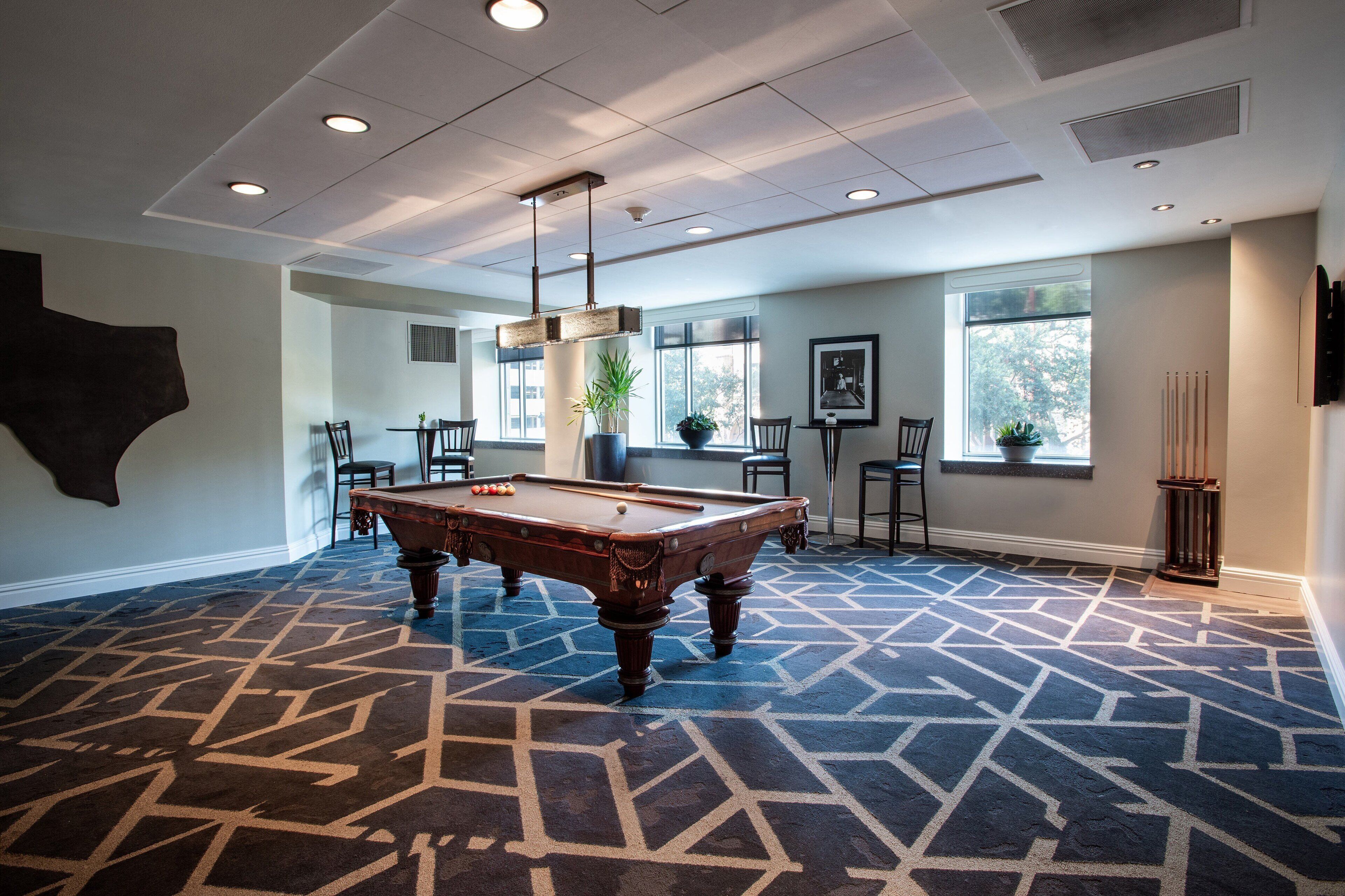 Une table de billard dans une pièce dotée d’un tapis à motifs, de chaises et de grandes fenêtres.