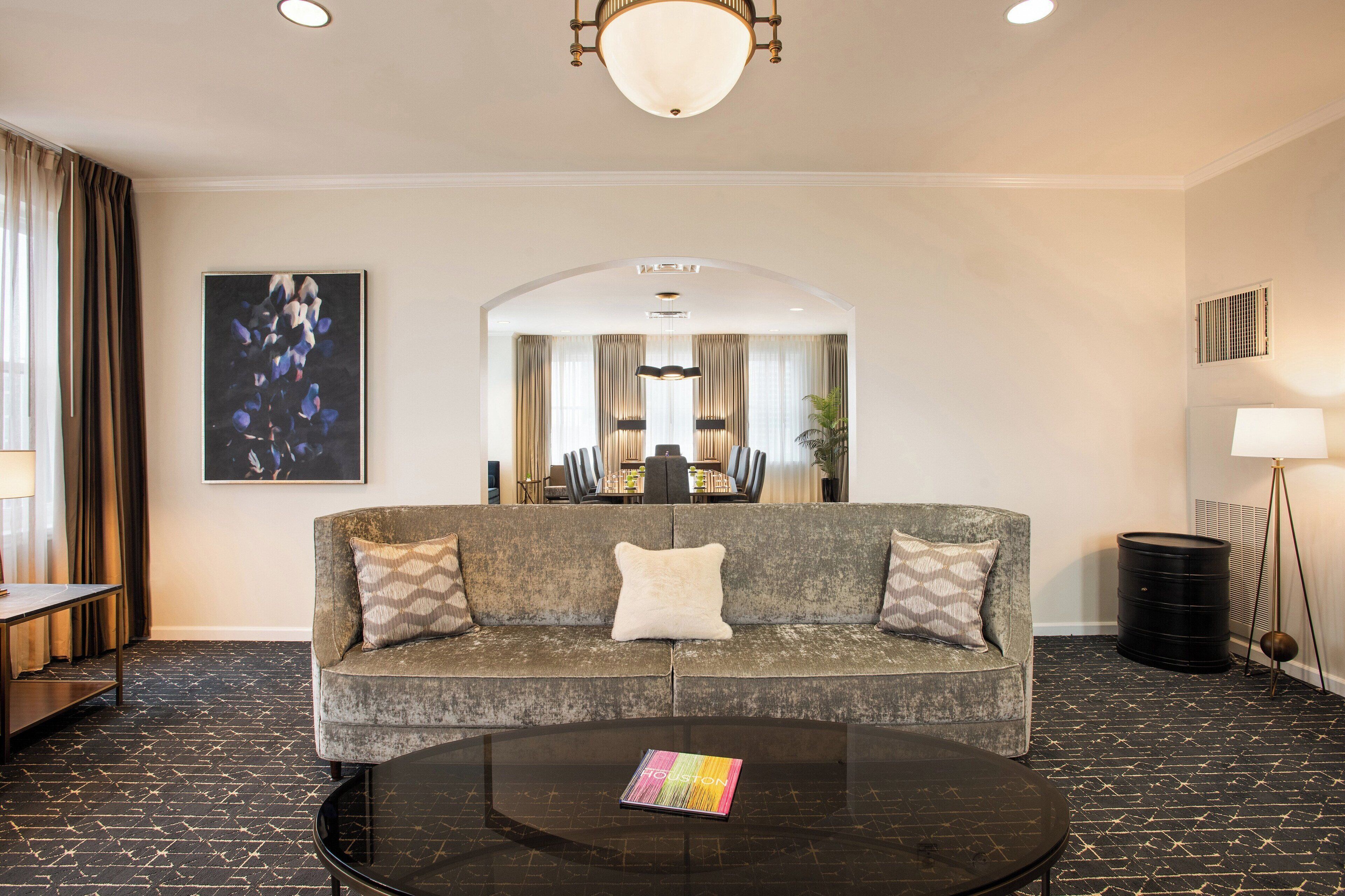 A modern hotel room with a sofa, a coffee table, a dining area in the background, and a large mirror.