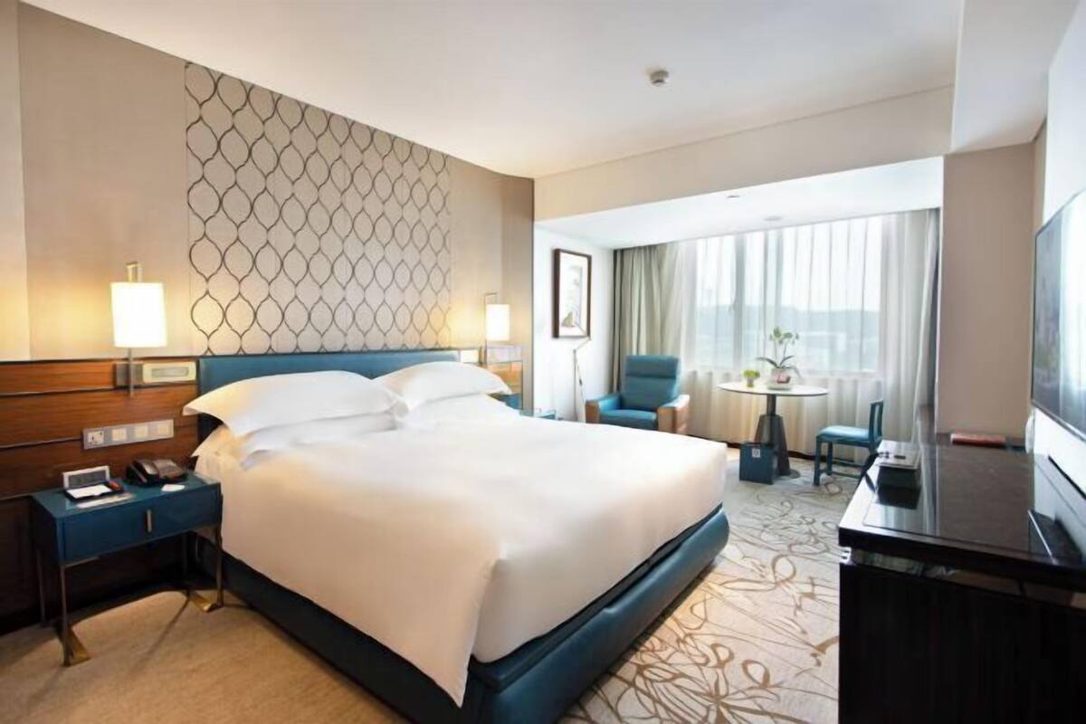 Una habitación de hotel con una cama grande, mesitas de noche, un escritorio con televisor, una pequeña mesa redonda con sillas y un amplio ventanal con cortinas.