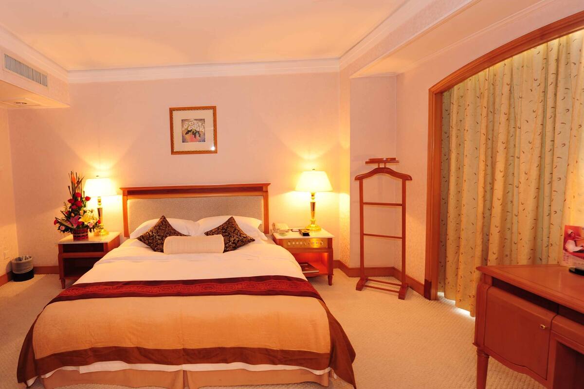 Habitación de hotel con una cama grande, dos mesitas de noche con lámparas, una silla de madera, un escritorio pequeño y un cuadro enmarcado en la pared.