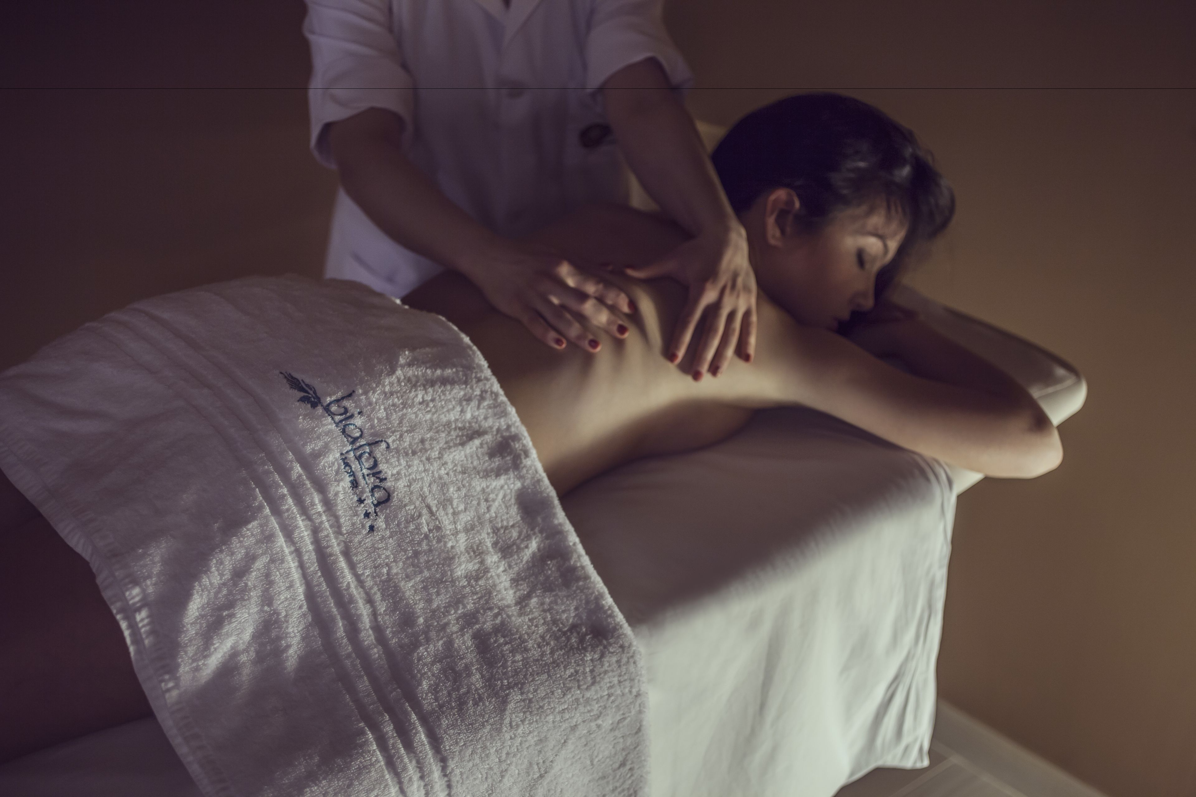 Sesión de masoterapia en una habitación con una cama cubierta con una sábana blanca y una toalla con texto.