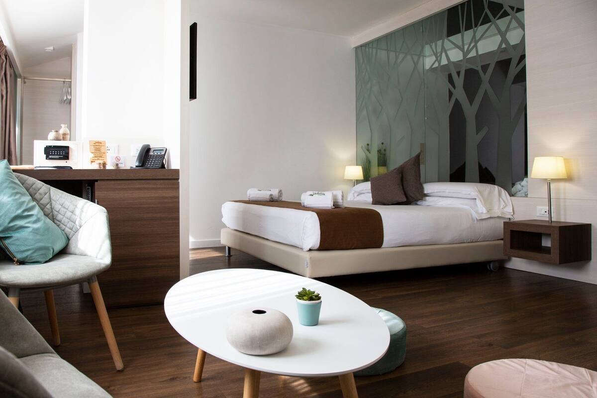 A modern hotel room with a bed, bedside table, chair, and a small table with a plant.