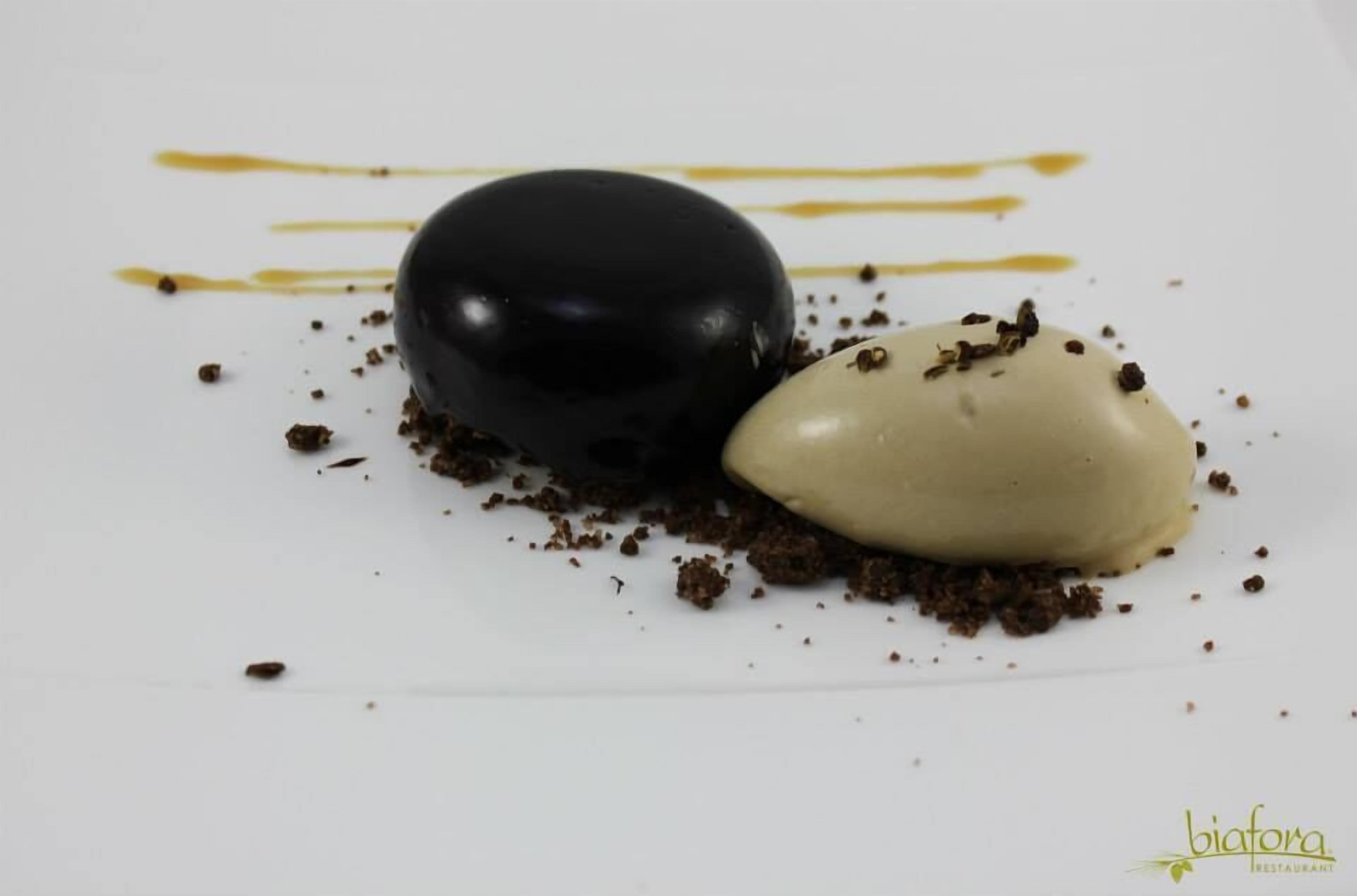 Un postre con una esfera de chocolate y una esfera blanca sobre un plato blanco con migas de chocolate y salsa amarilla.