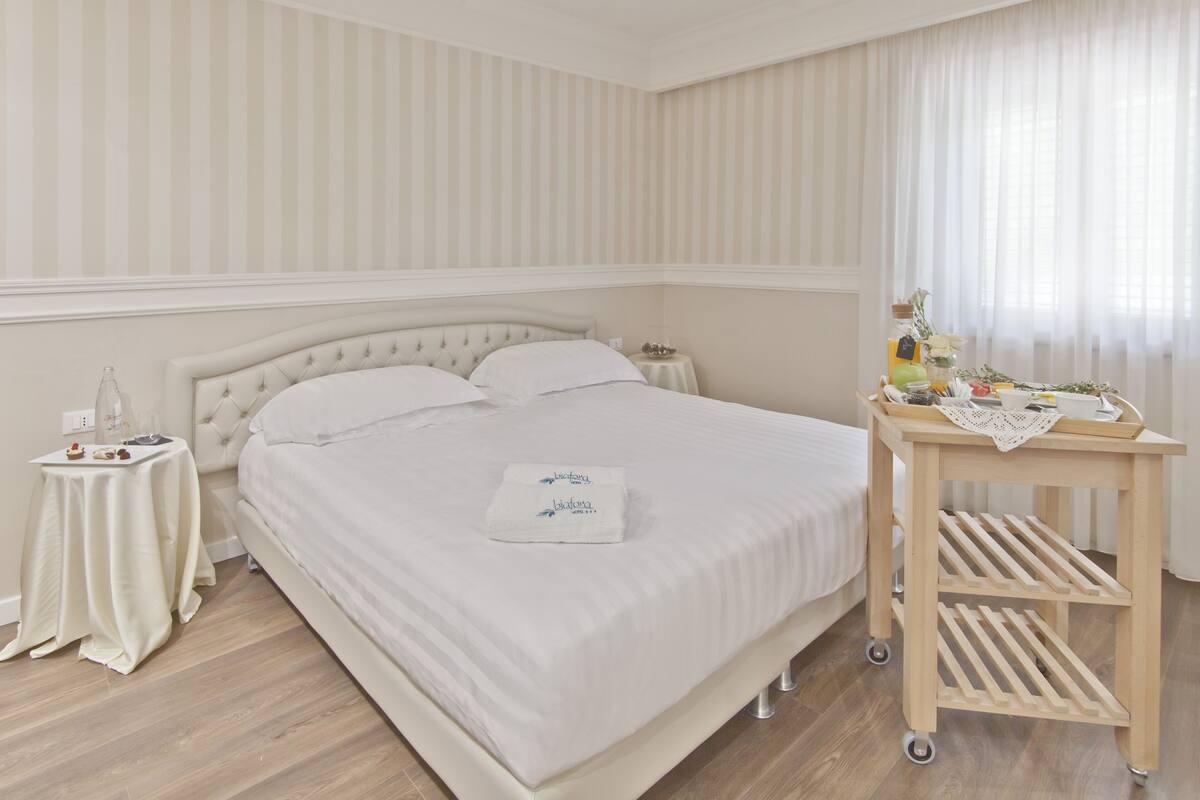 A neatly made bed with a white bedspread and pillows, a bedside table with a bottle and a glass, and a wooden cart with a tray.