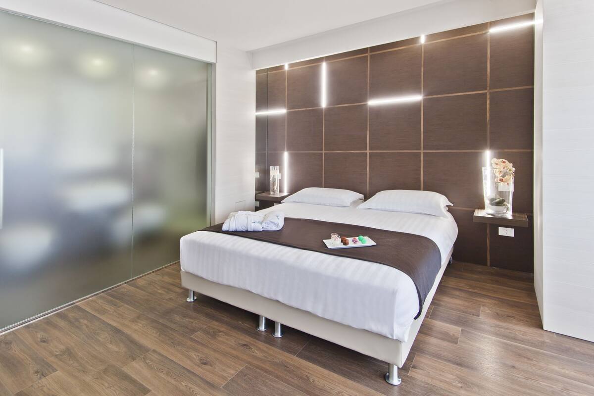 A modern hotel room with a large bed, wooden flooring, a glass partition wall, and a decorative wall panel.