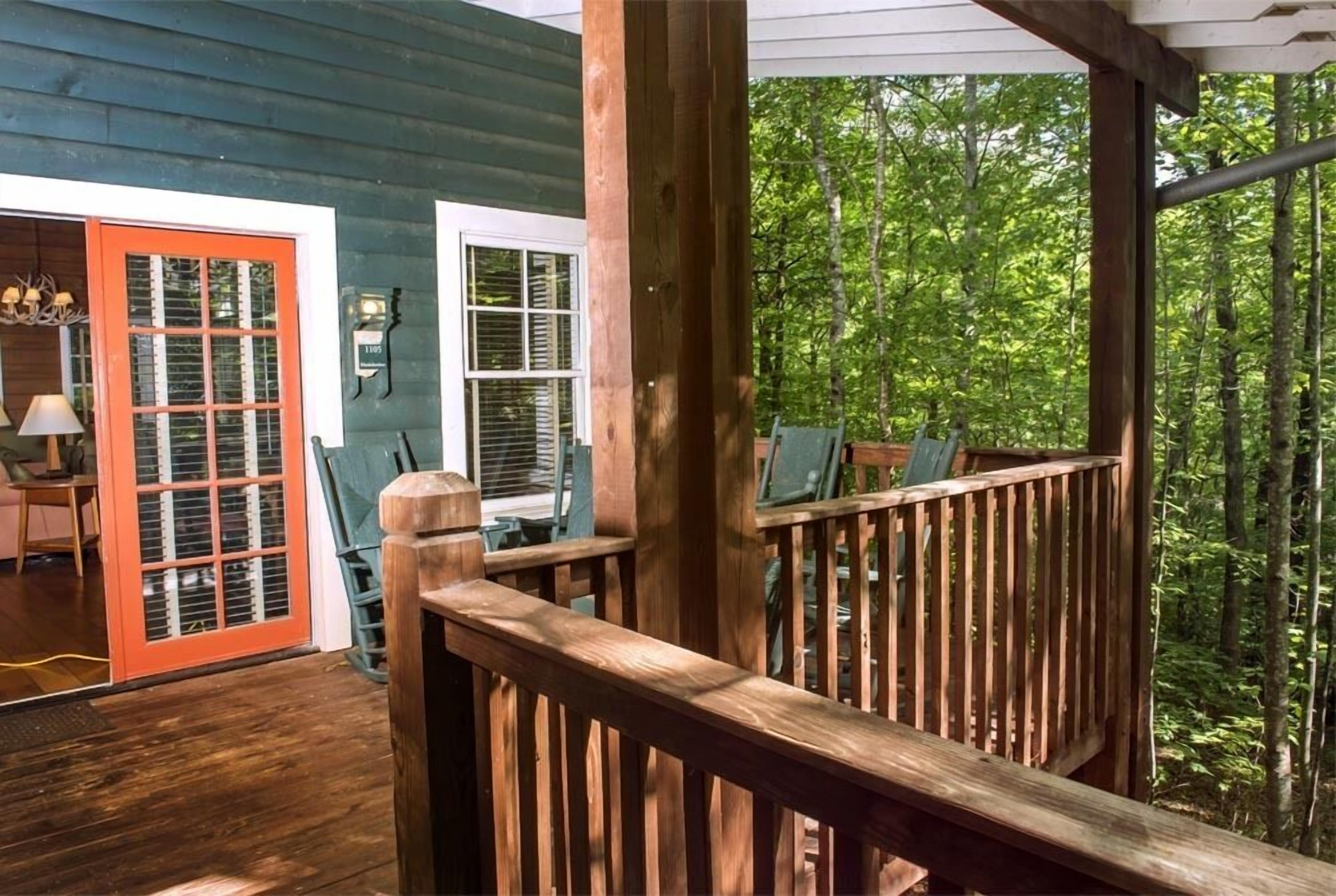 A wooden deck with a railing, a door with a red frame, and a view of a forested area.