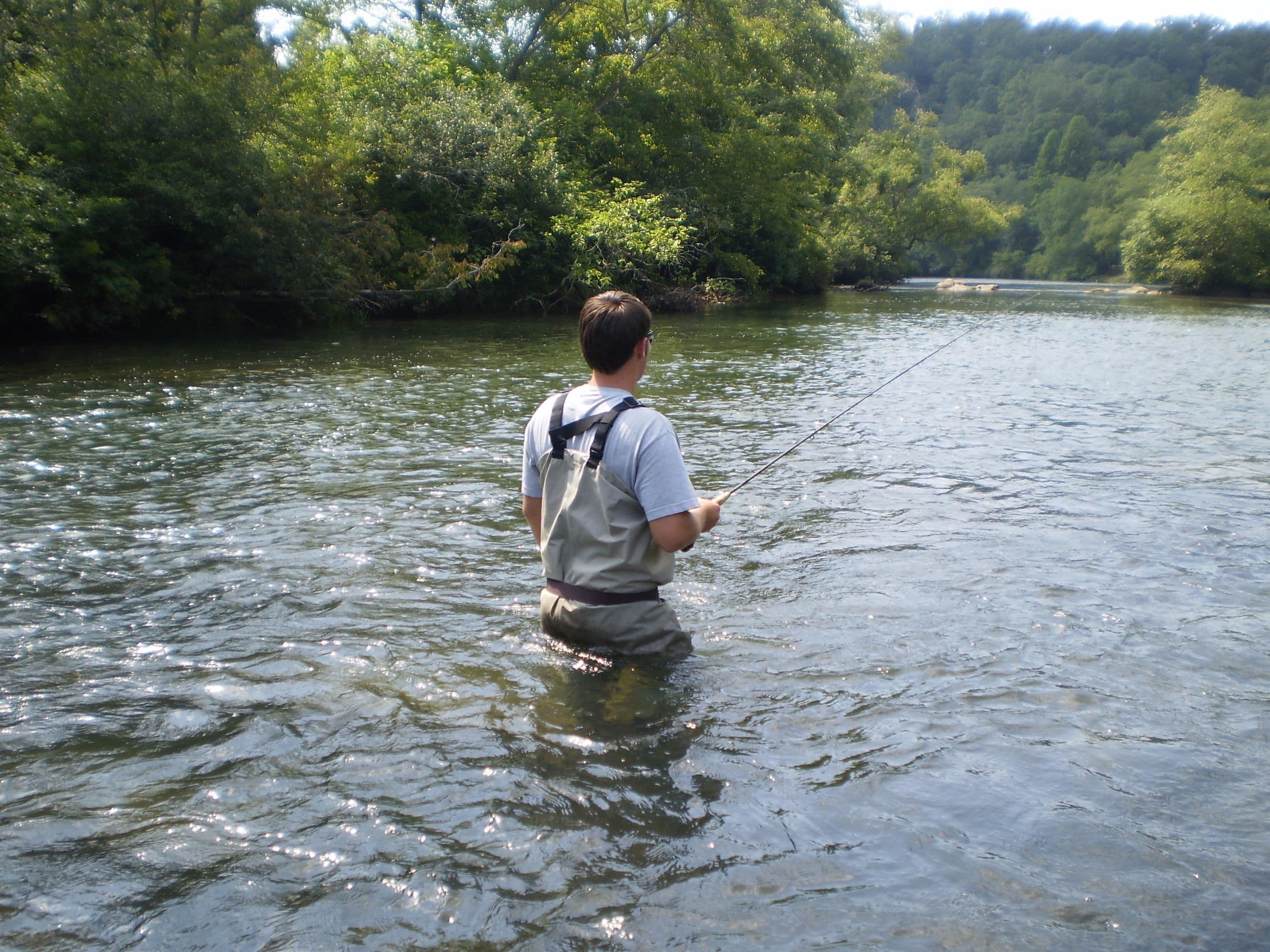 A person standing in a river, wearing a light-colored shirt and a vest, holding a fishing rod.