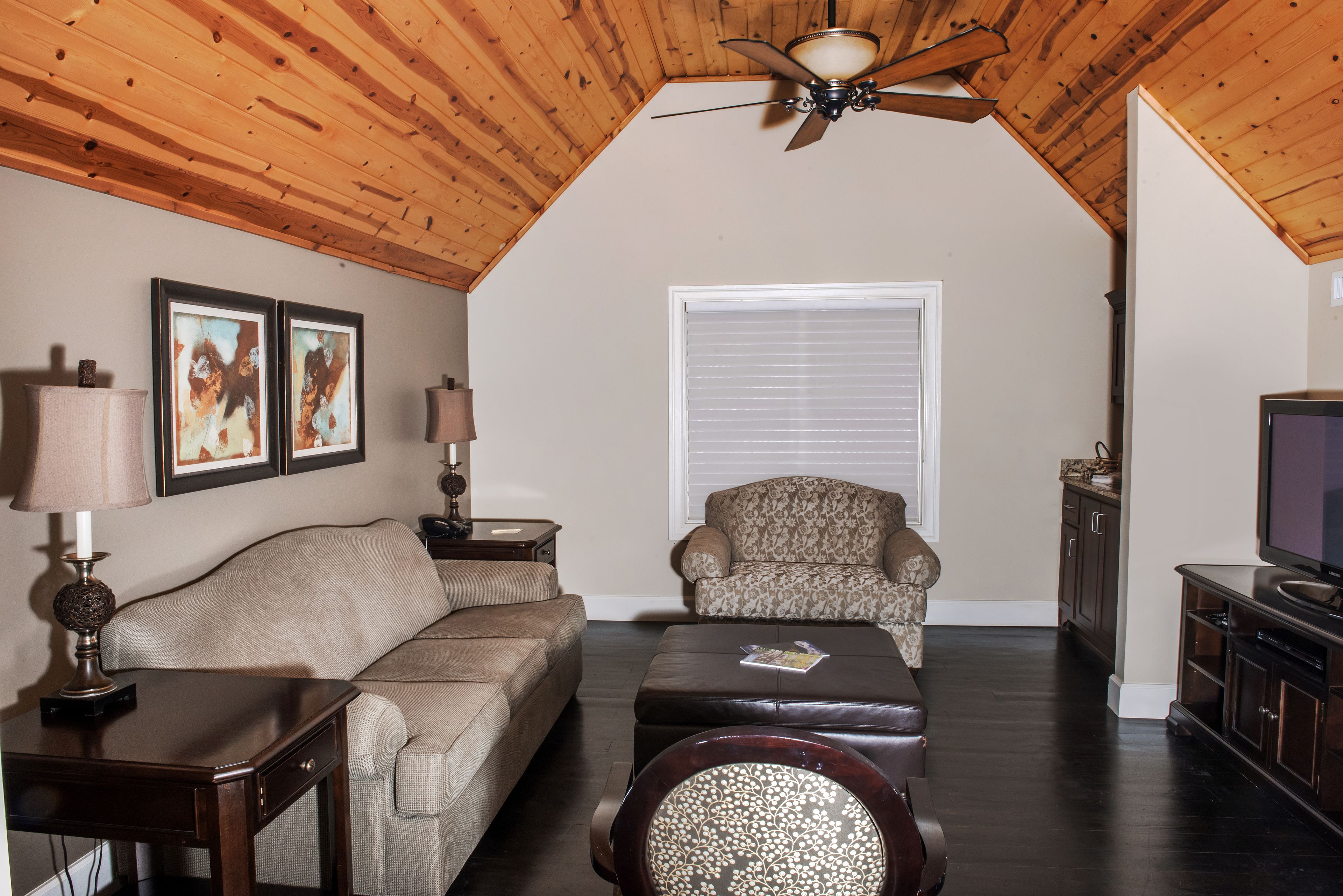A living room with a wooden ceiling, a sofa, a patterned armchair, a coffee table, a television, and framed artwork on the wall.