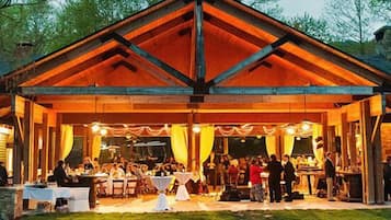 A covered outdoor dining area with multiple tables and chairs, surrounded by wooden beams and a pitched roof.