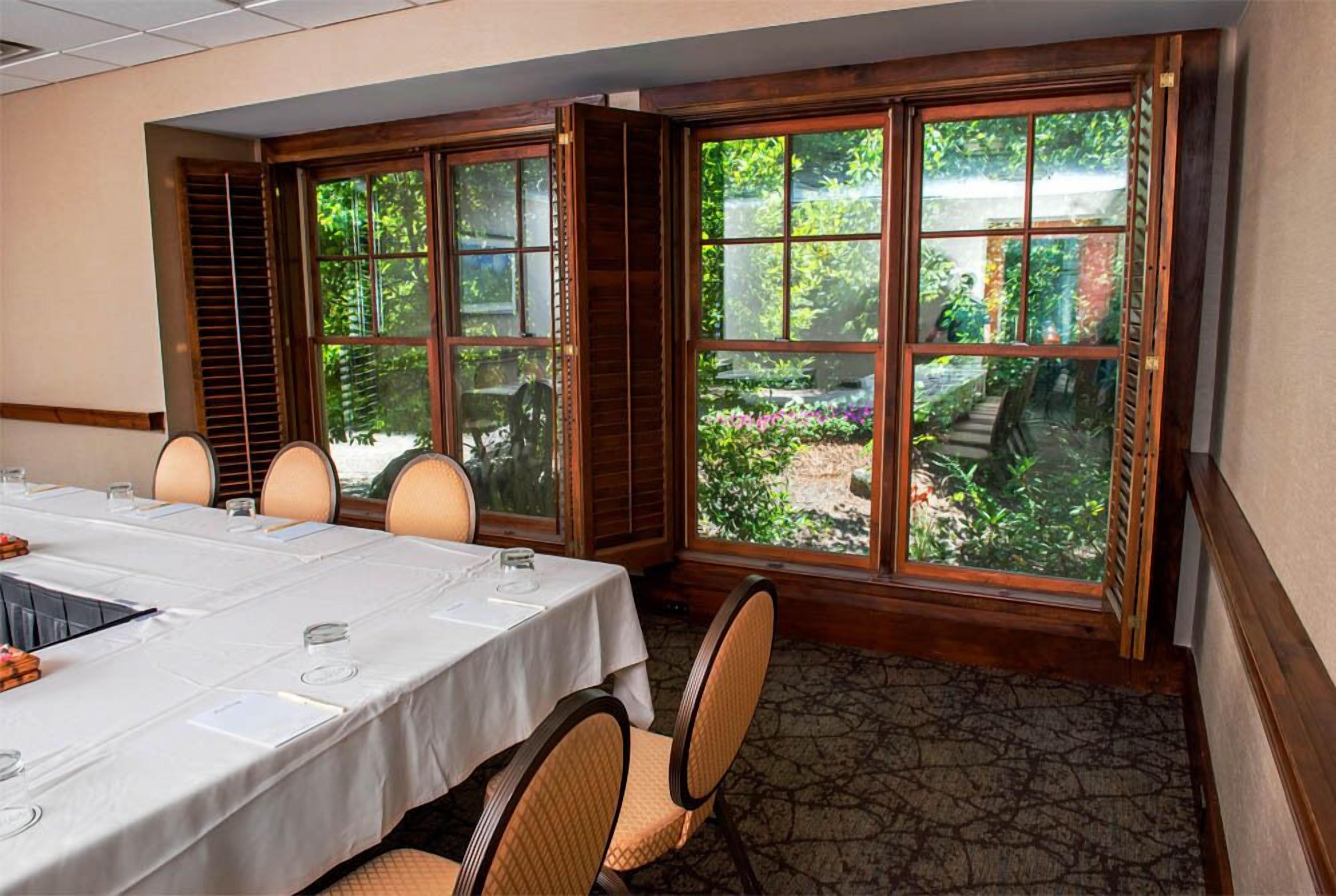 A conference room with a long table, chairs, and large windows offering a view of a garden.