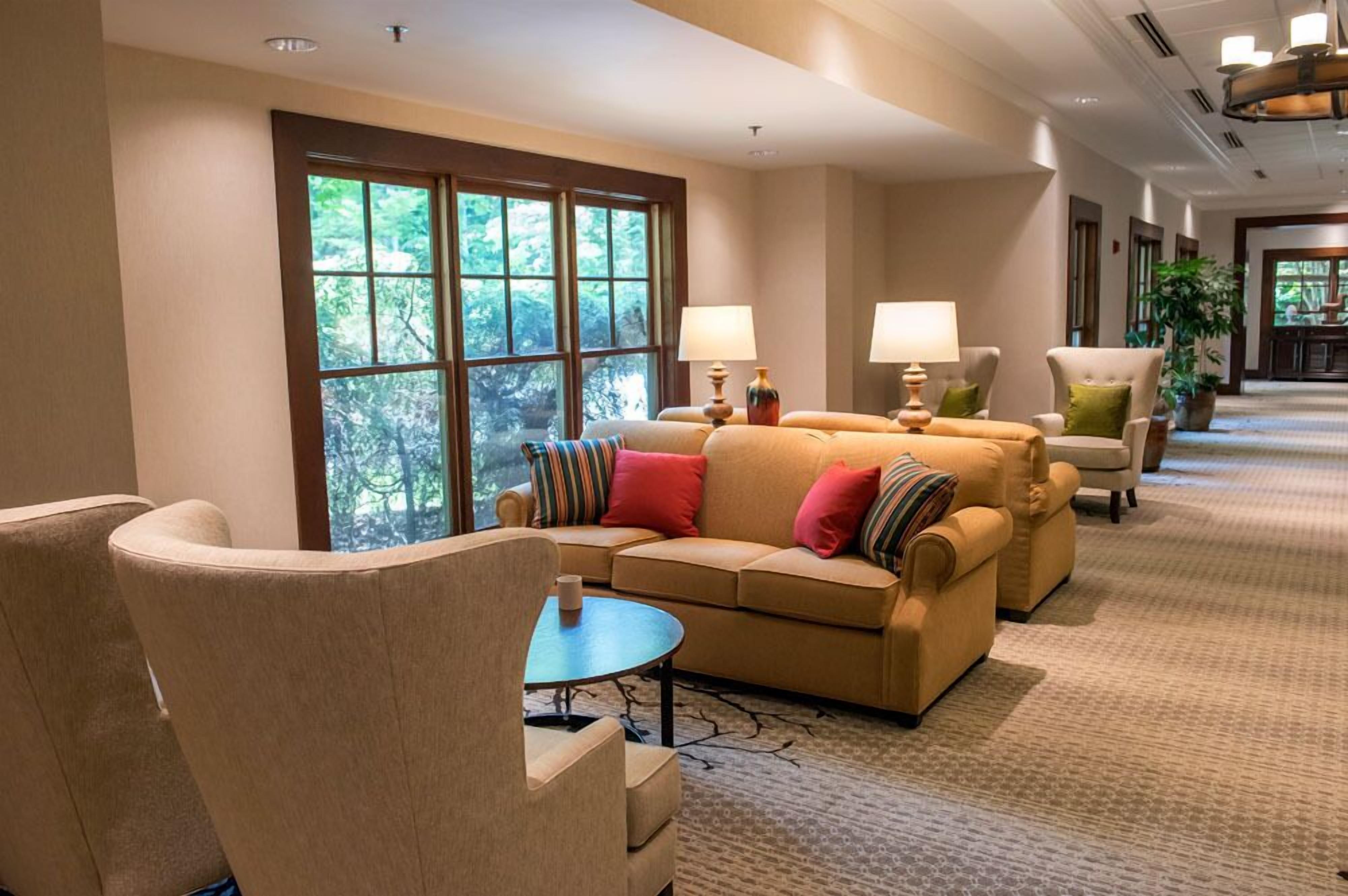 A spacious lobby with a beige sofa, armchairs, and a coffee table. Large windows provide a view of greenery outside.