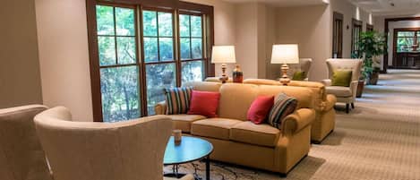 A spacious lobby with a beige sofa, armchairs, and a coffee table. Large windows provide a view of greenery outside.