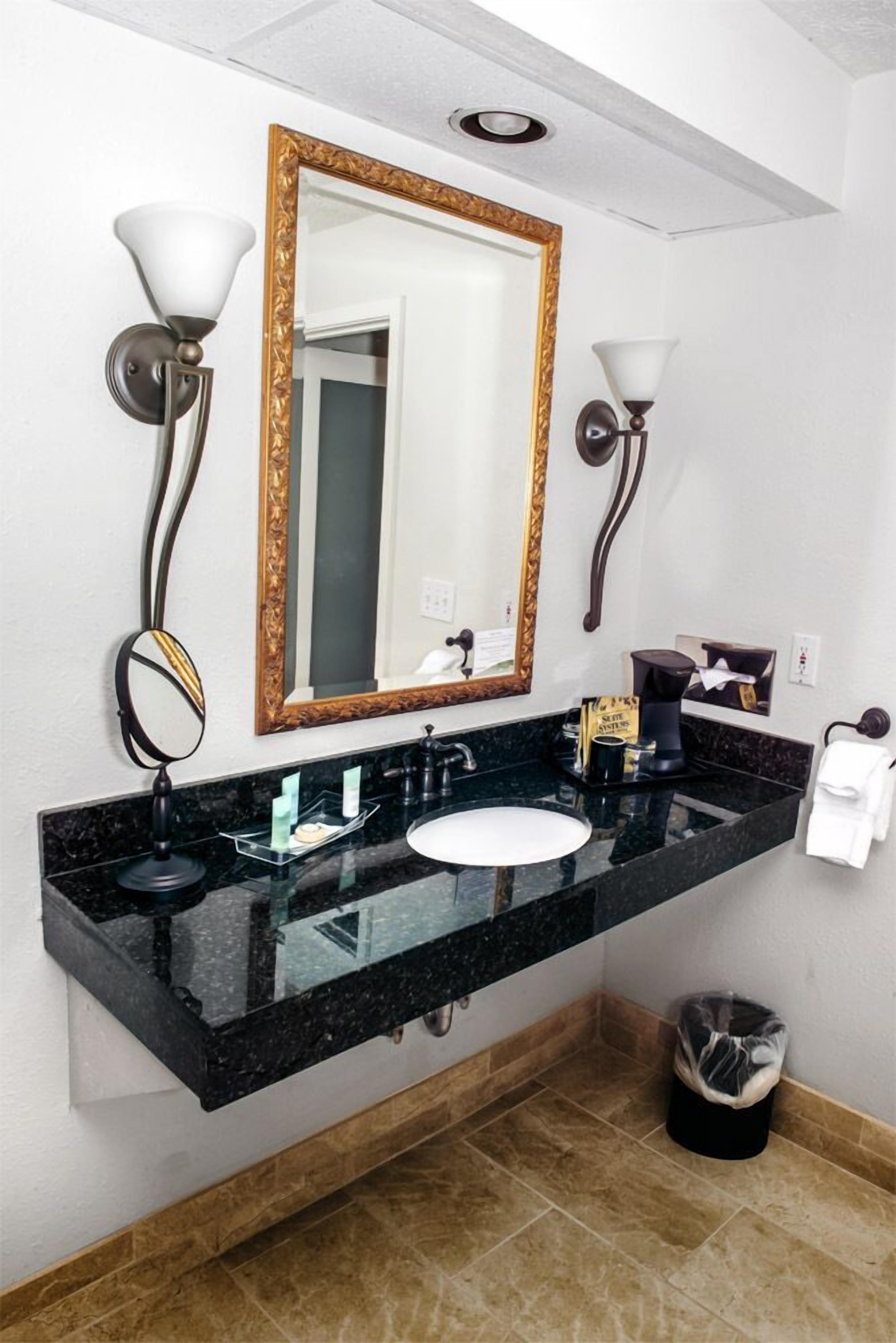 Une salle de bain avec un lavabo en marbre noir, un miroir encadré d’or et deux lampes fixées au mur.