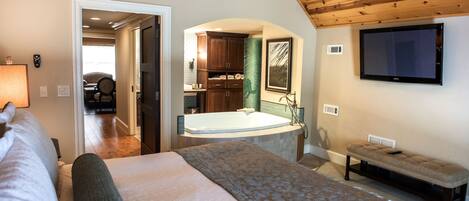 Un dormitorio con una cama grande, una banca, un televisor y un baño con bañera y ducha.