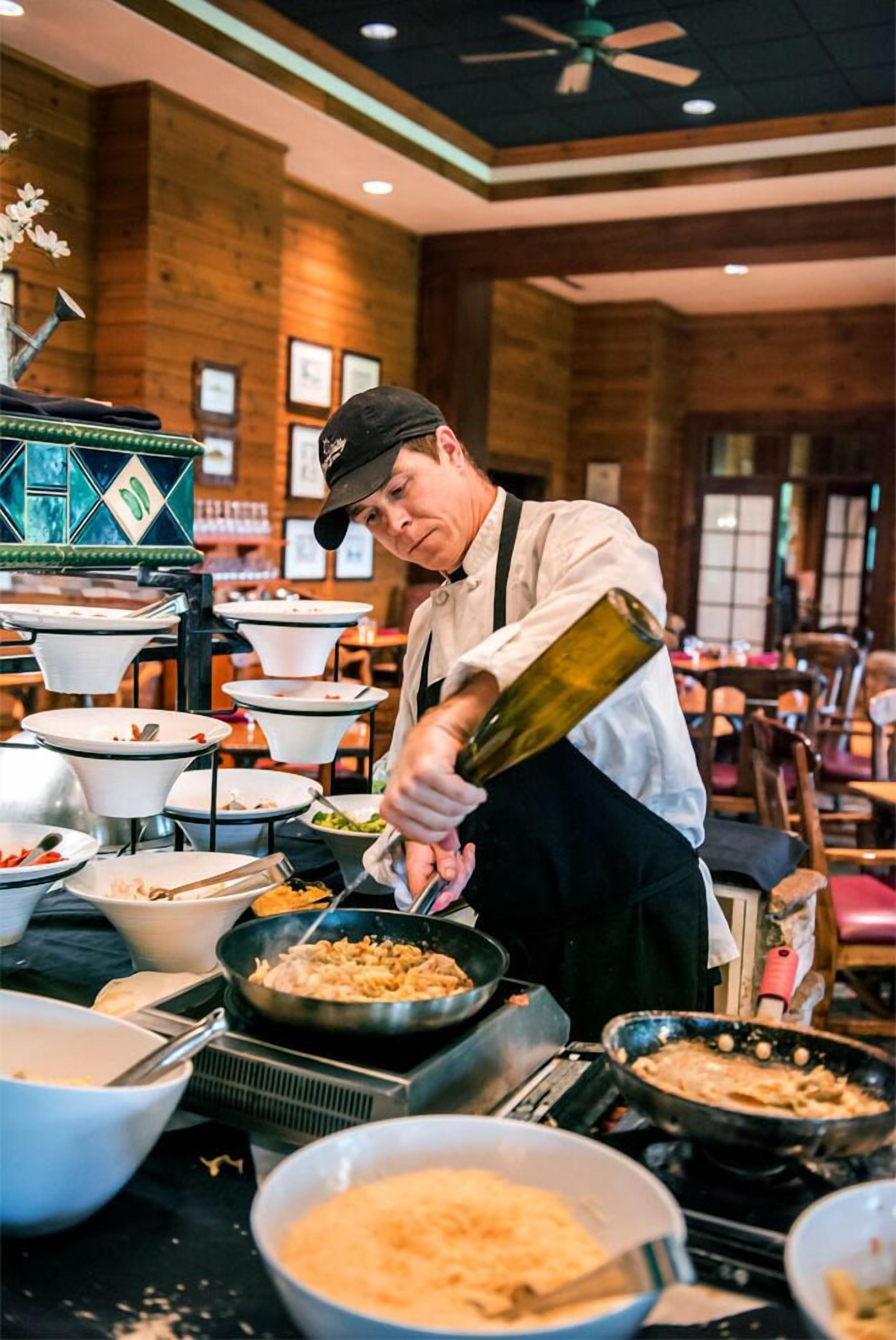 A chef preparing food in a kitchen with multiple cooking stations and a buffet setup.