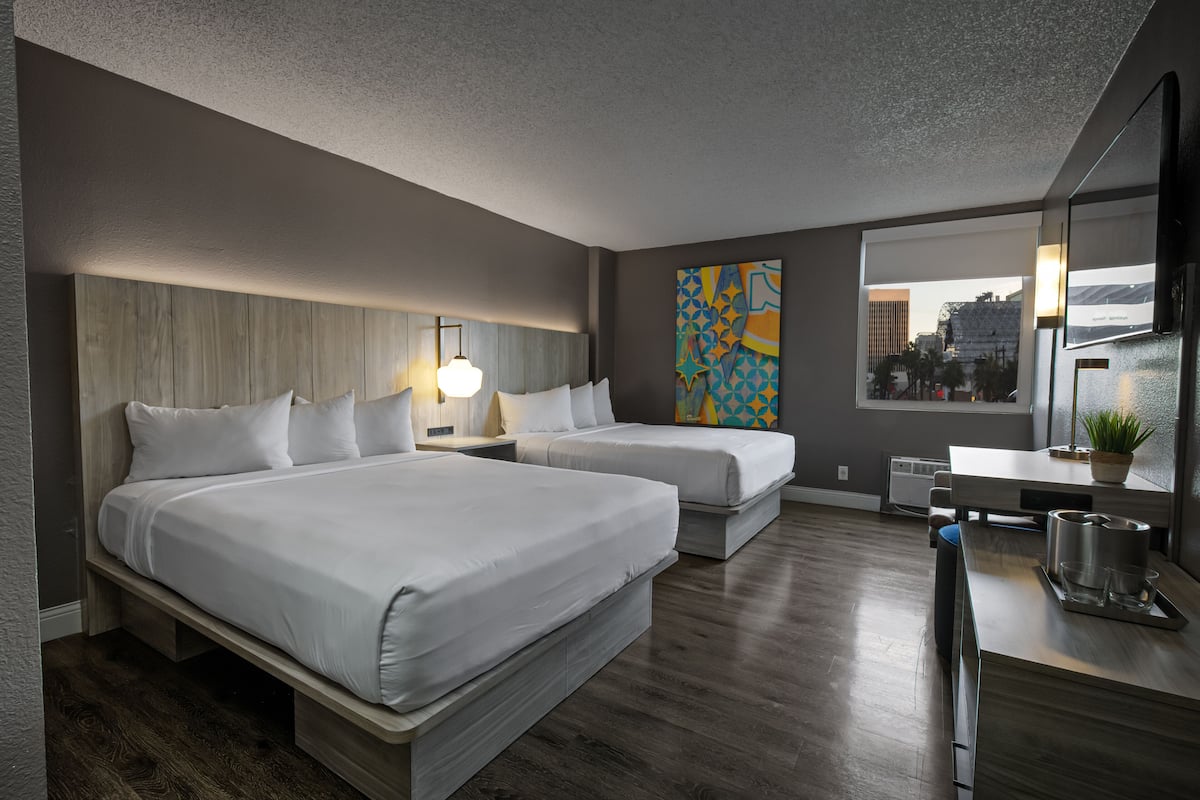 A modern hotel room with two beds, a desk, a large window with a city view, and a decorative wall piece.